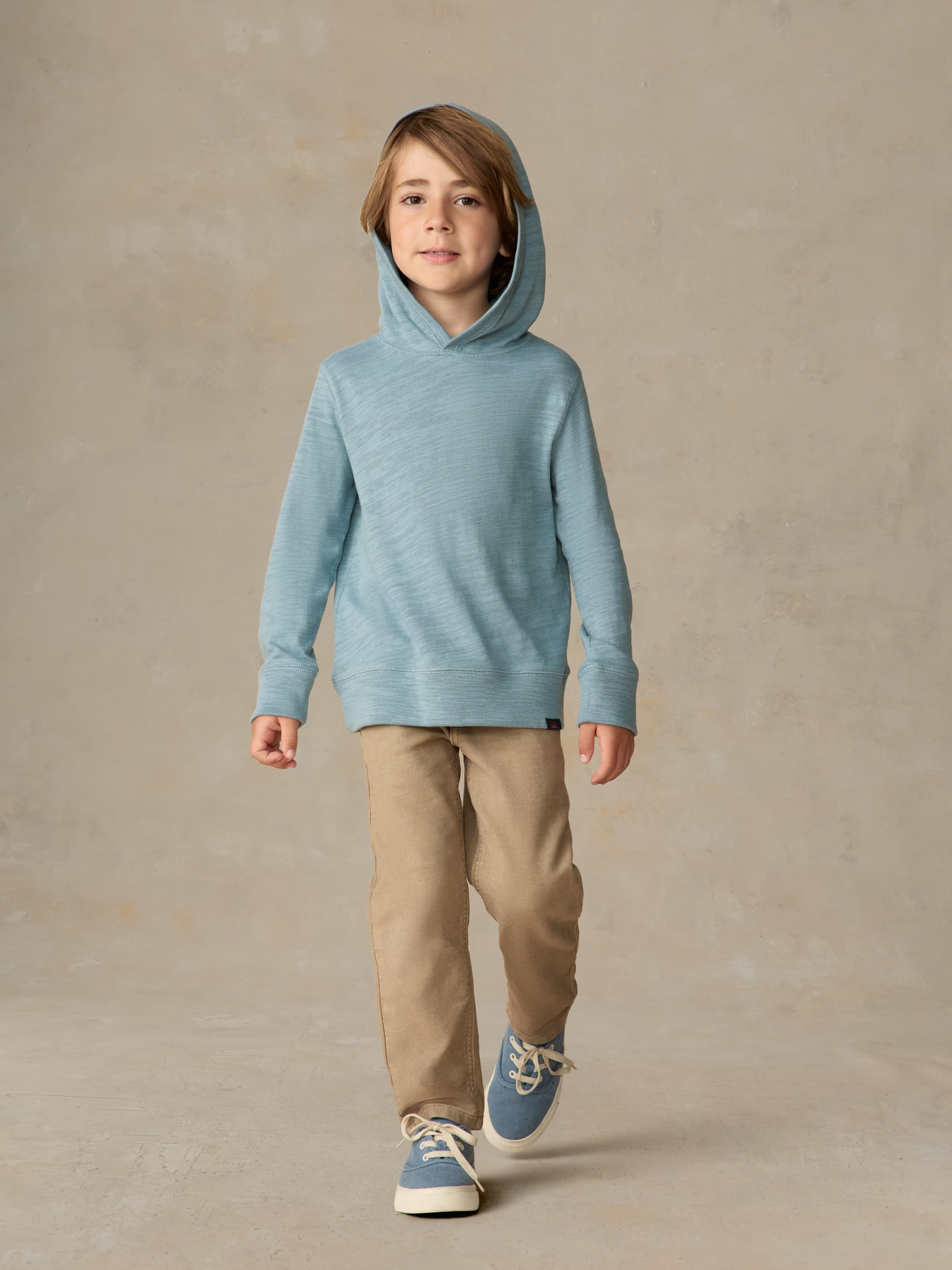 Child wearing a light blue hoodie and beige pants against a beige background