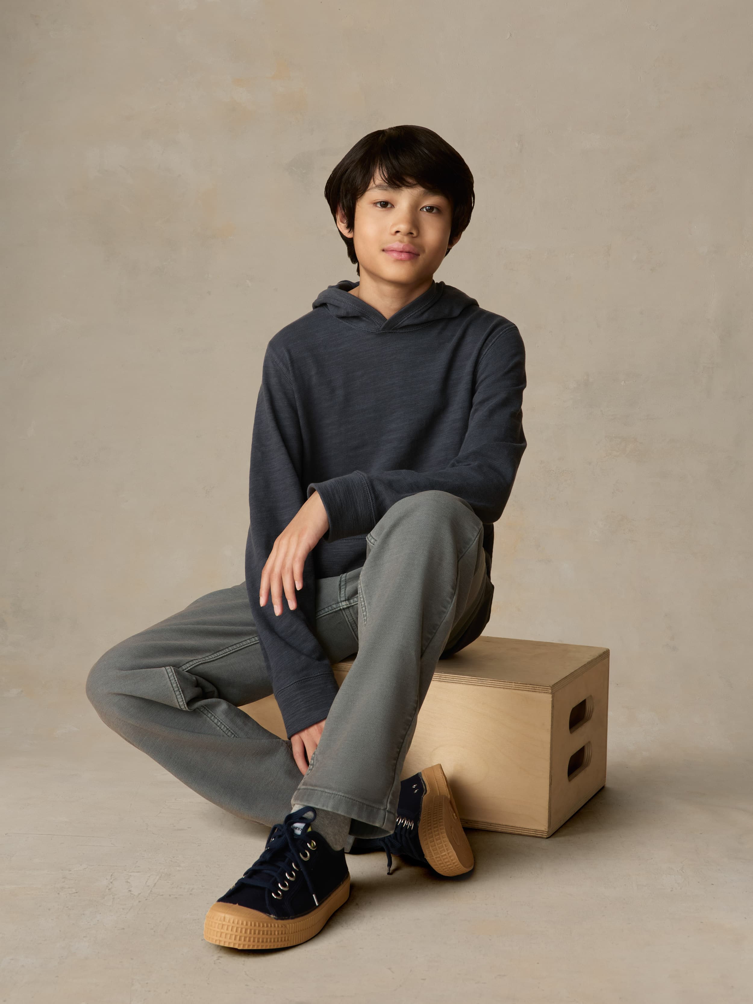 Child sitting on a wooden box wearing a navy hoodie and grey pants against a beige background
