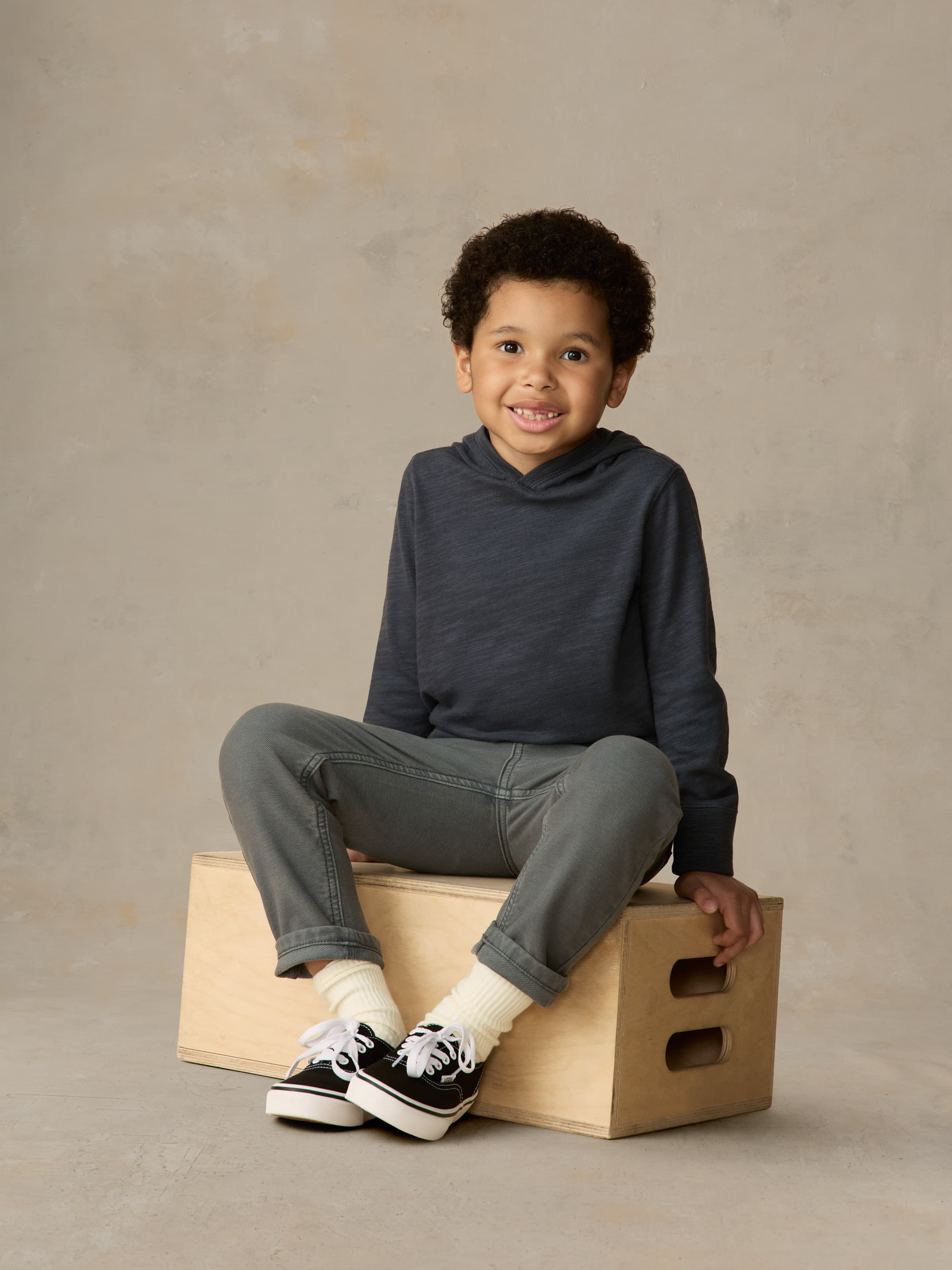 Child sitting on a wooden box wearing a navy hoodie and grey pants against a beige background