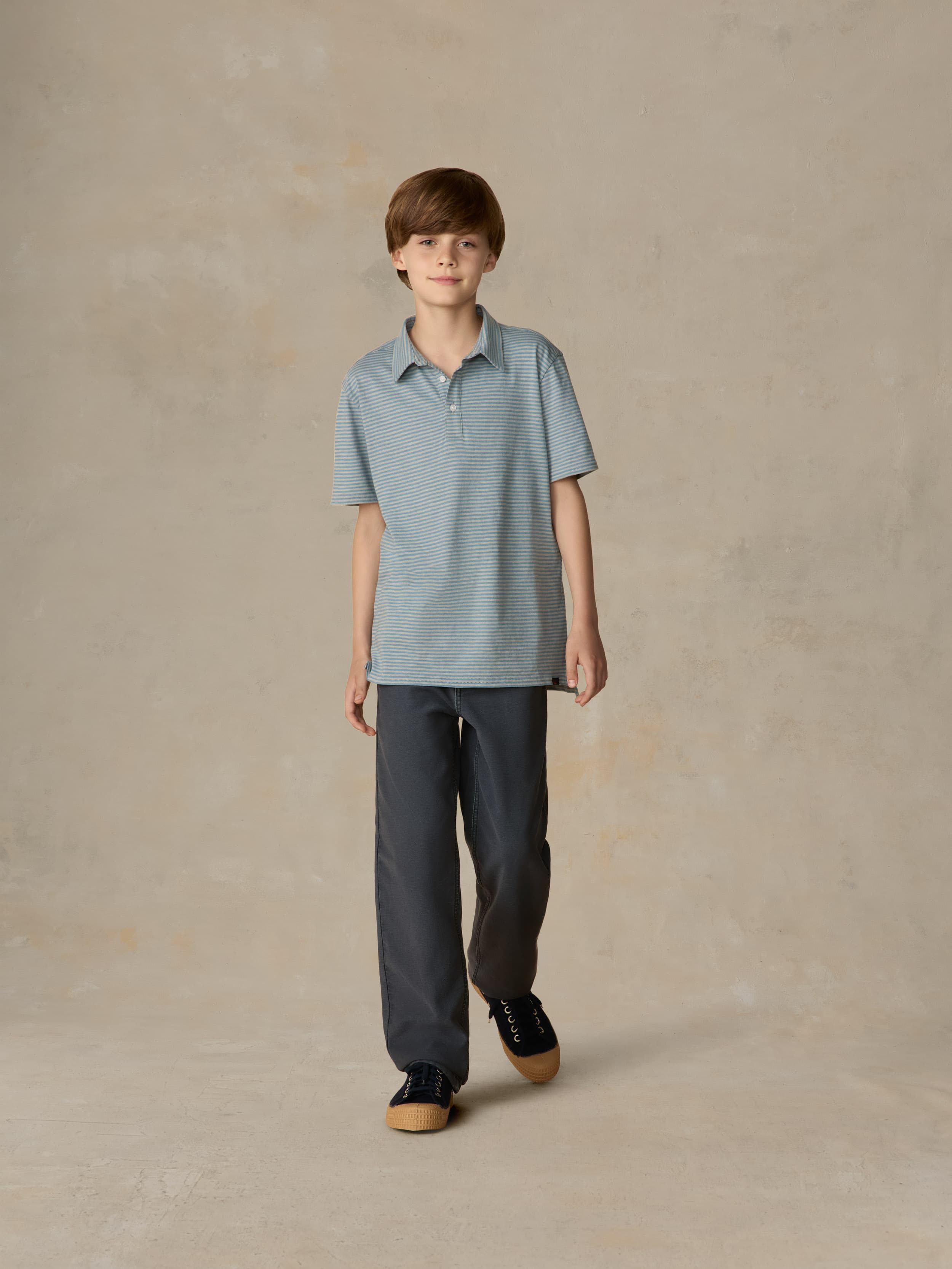 Child wearing a blue striped polo and navy pants against a beige background