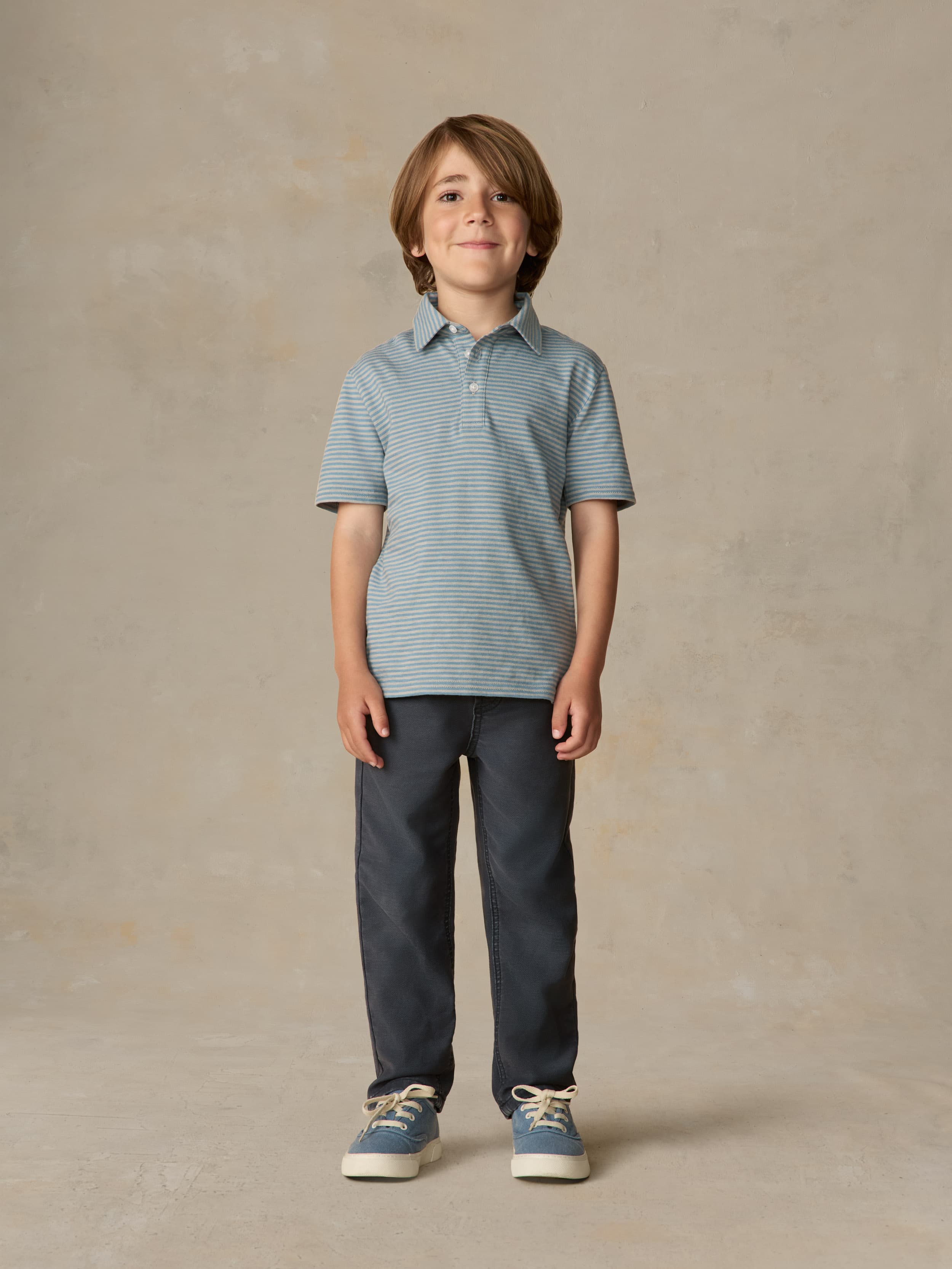 Child wearing a blue striped polo and navy pants against a beige background