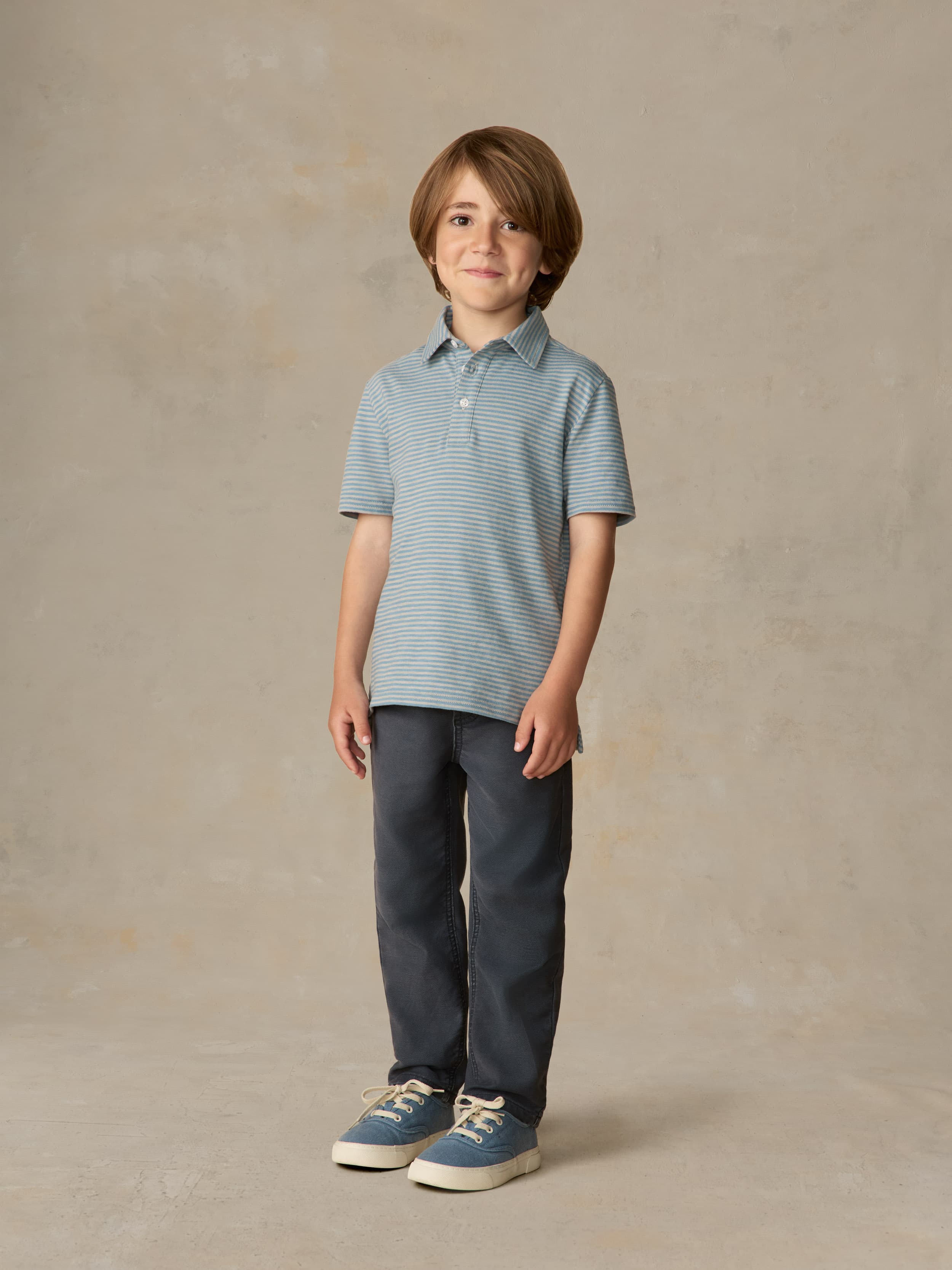 Child wearing a blue striped polo and navy pants against a beige background