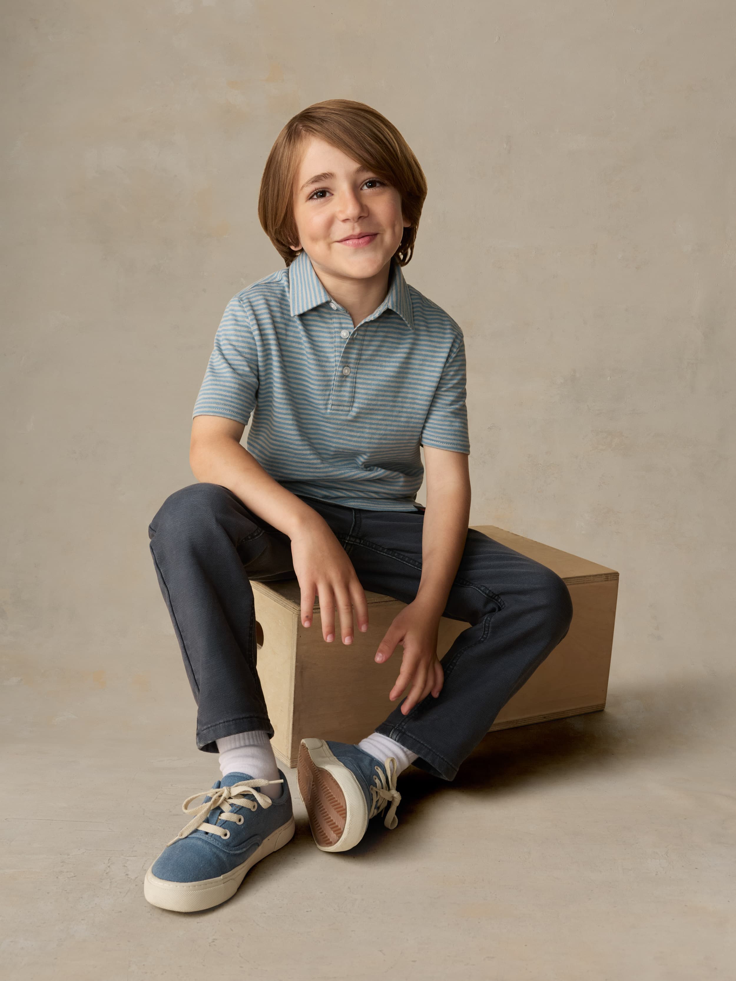 Child sitting on a wooden box wearing a blue striped polo and navy pants against a beige background
