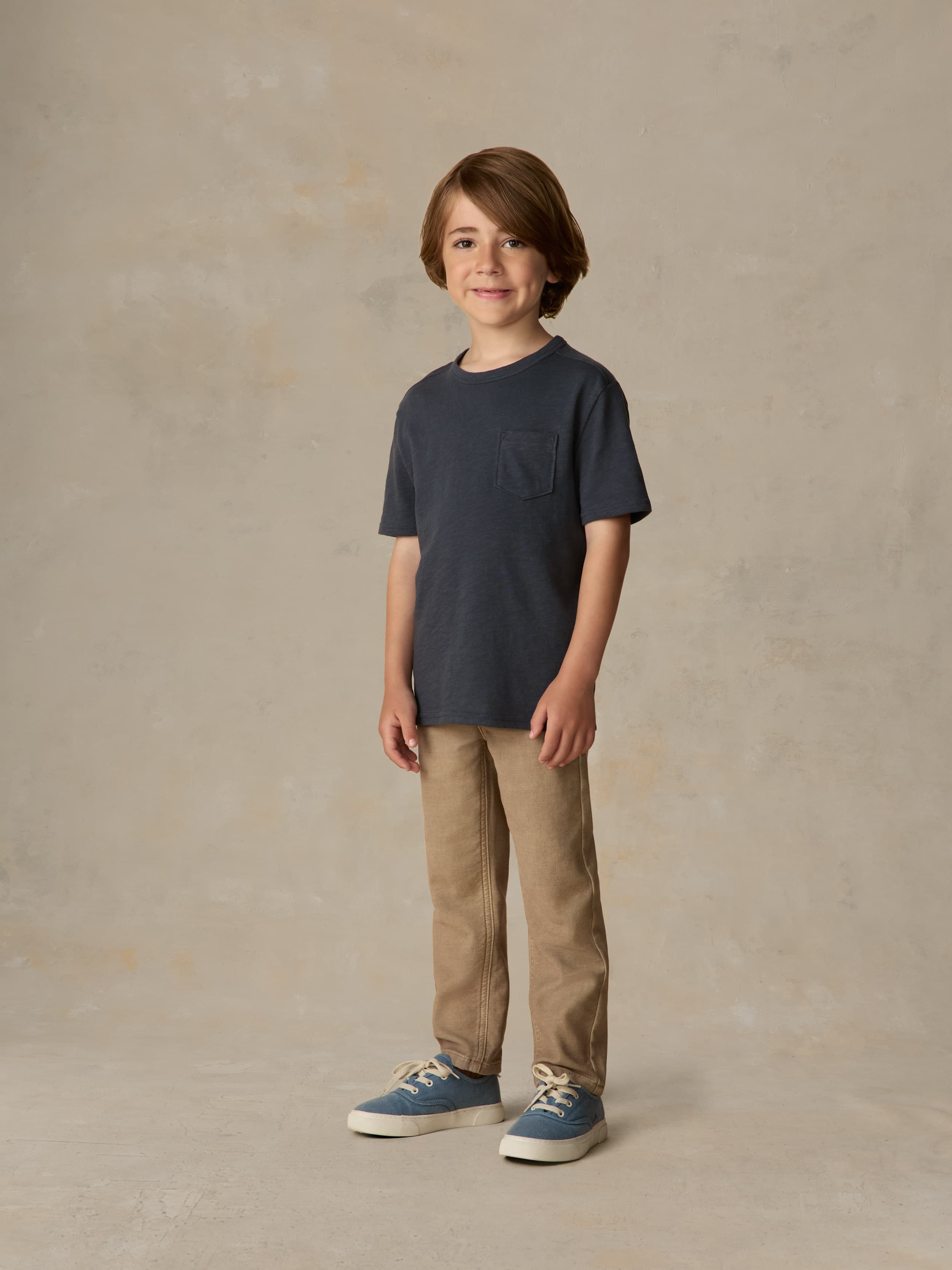 Child wearing a blue pocket tee and beige pants against a beige background