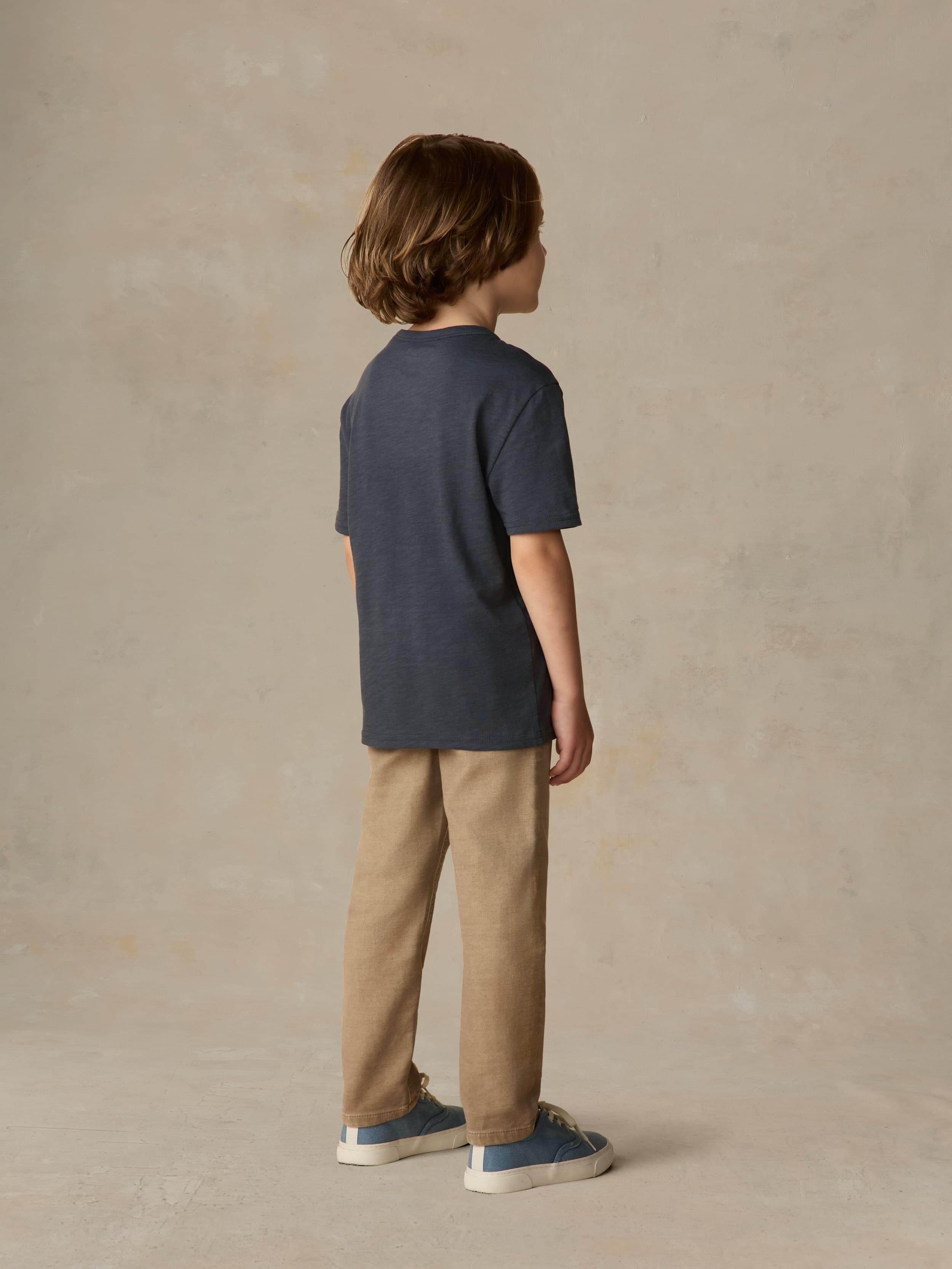 Child wearing a blue pocket tee and beige pants against a beige background
