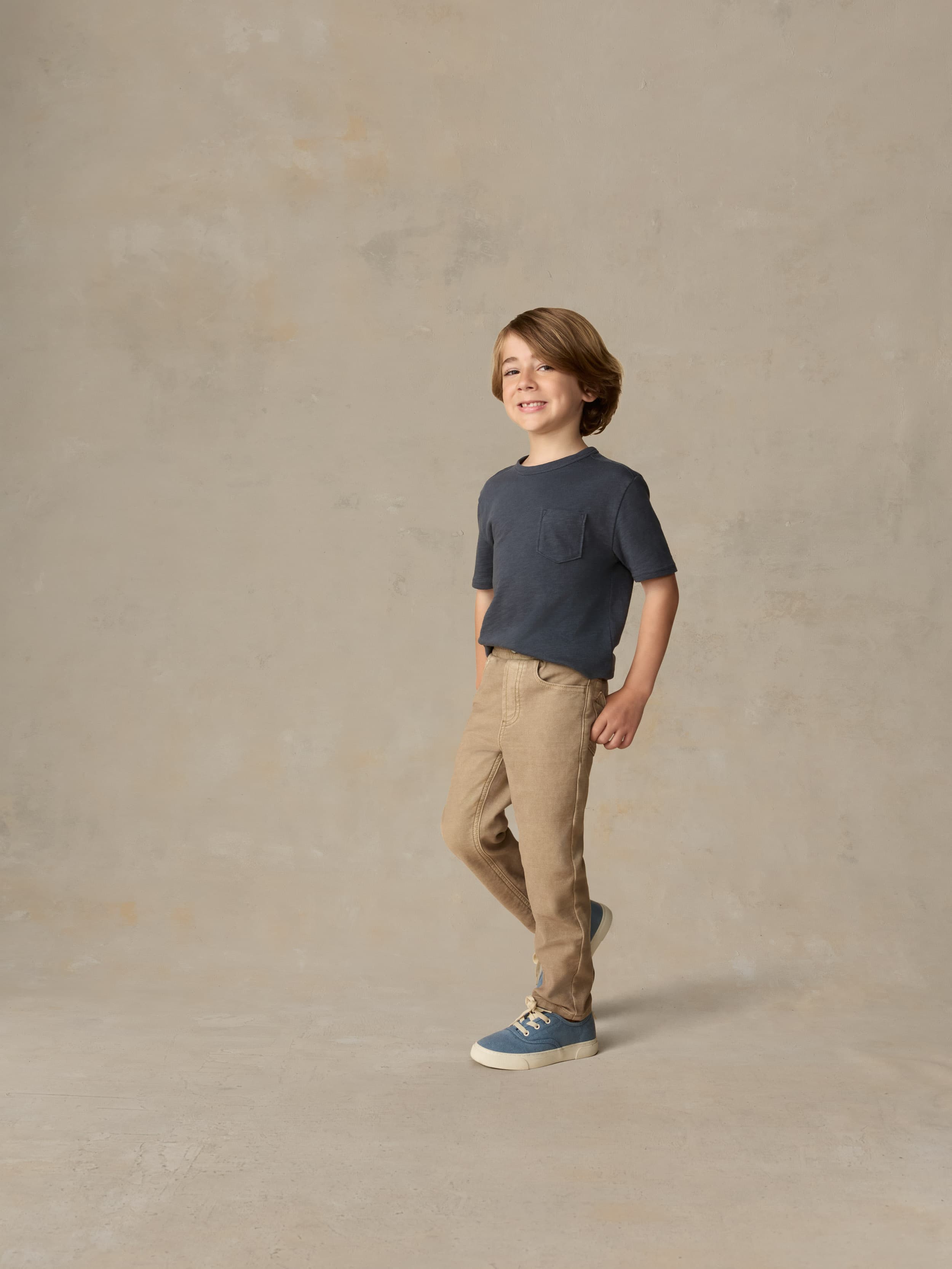 Child wearing a blue pocket tee and beige pants against a beige background