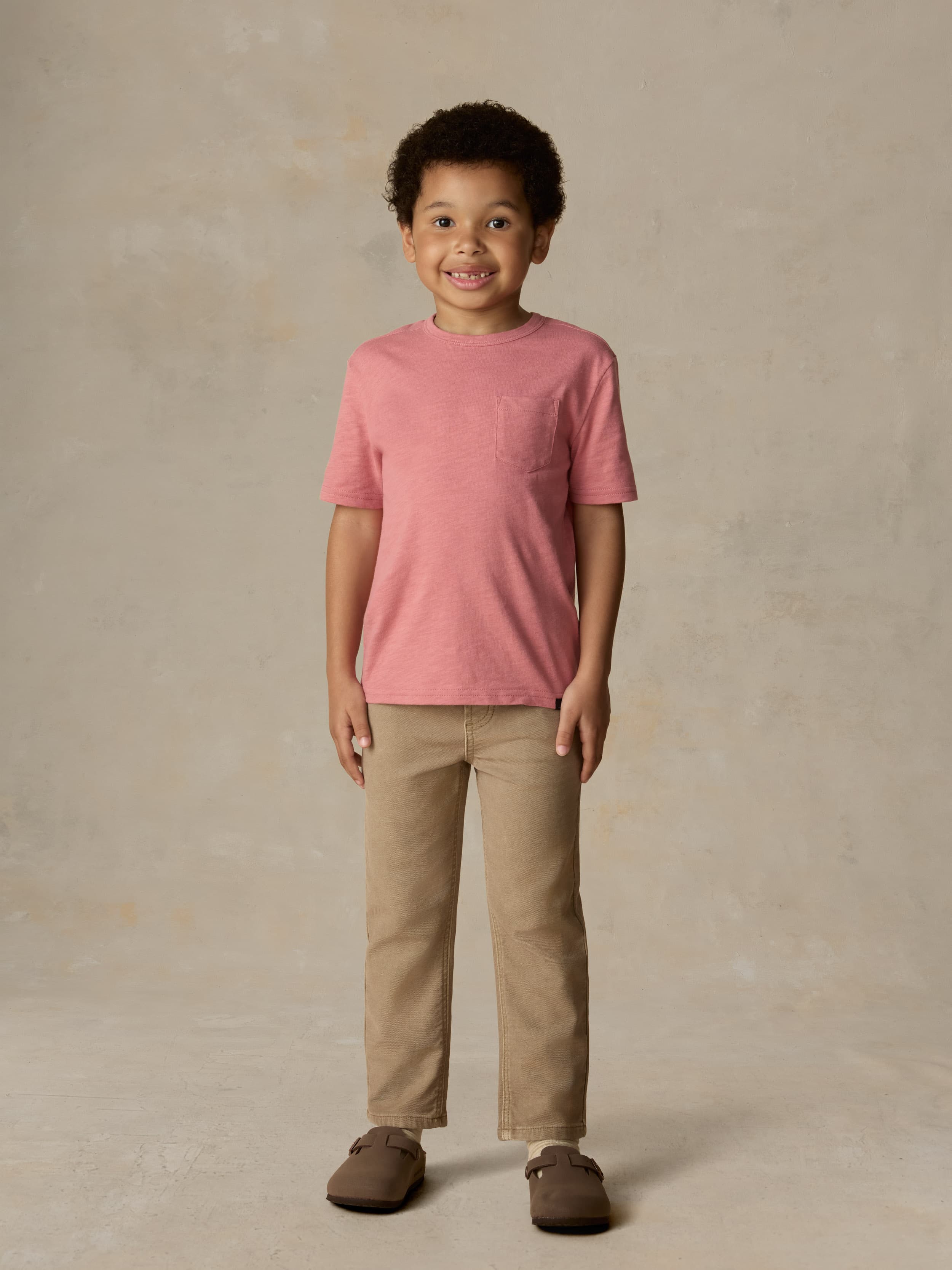 Child wearing a red pocket tee and beige pants against a beige background