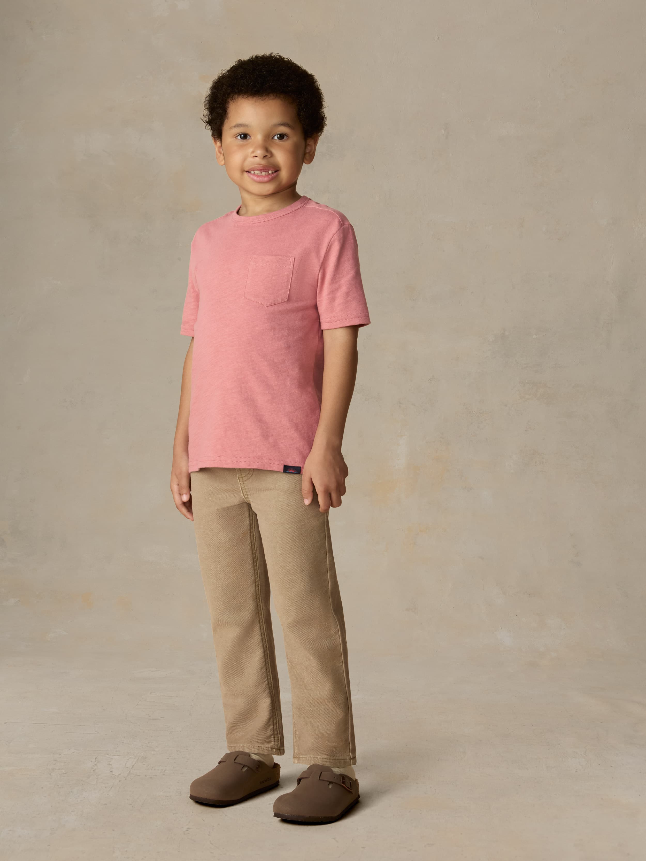 Child wearing a red pocket tee and beige pants against a beige background