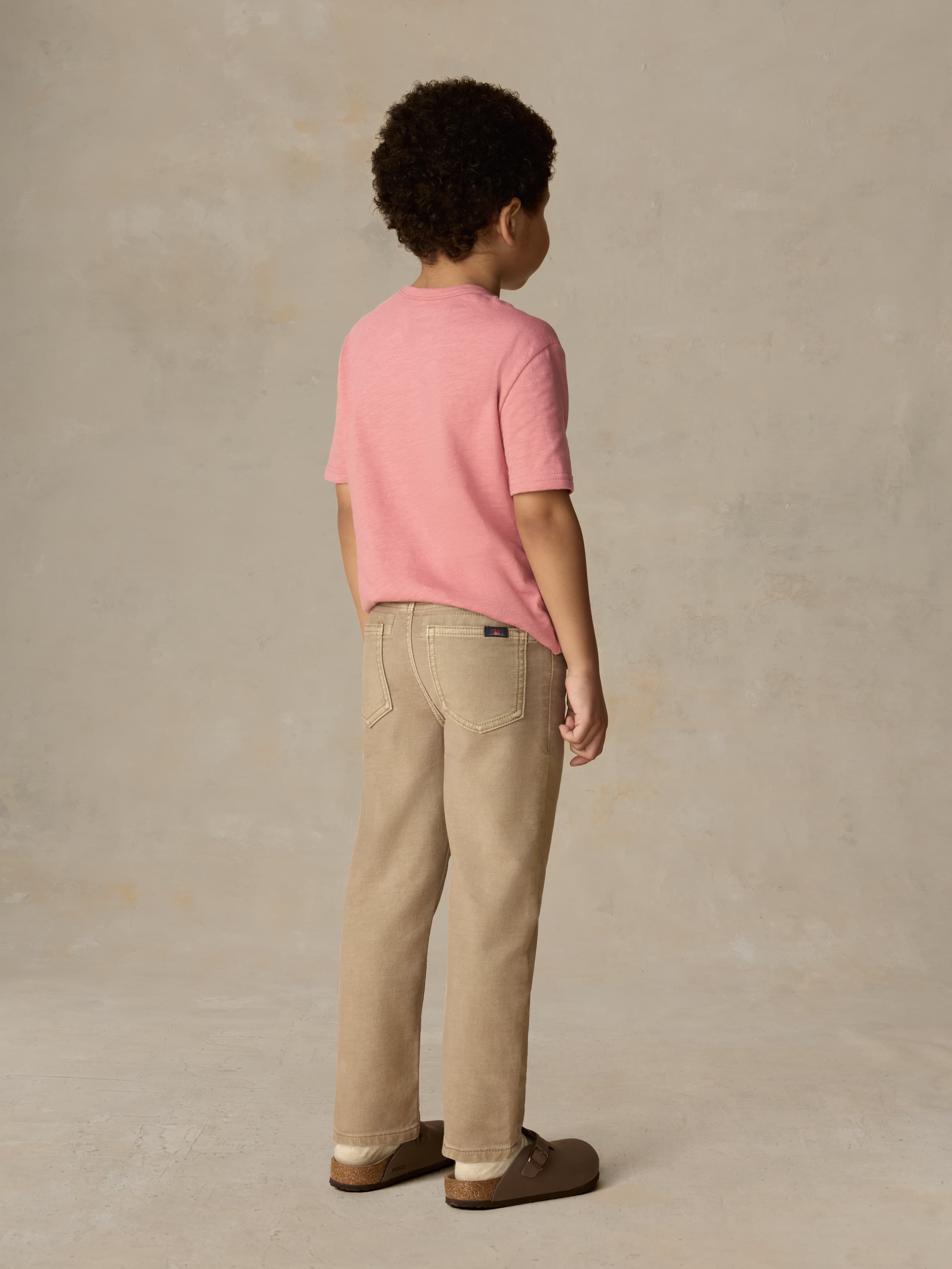 Child wearing a red pocket tee and beige pants against a beige background