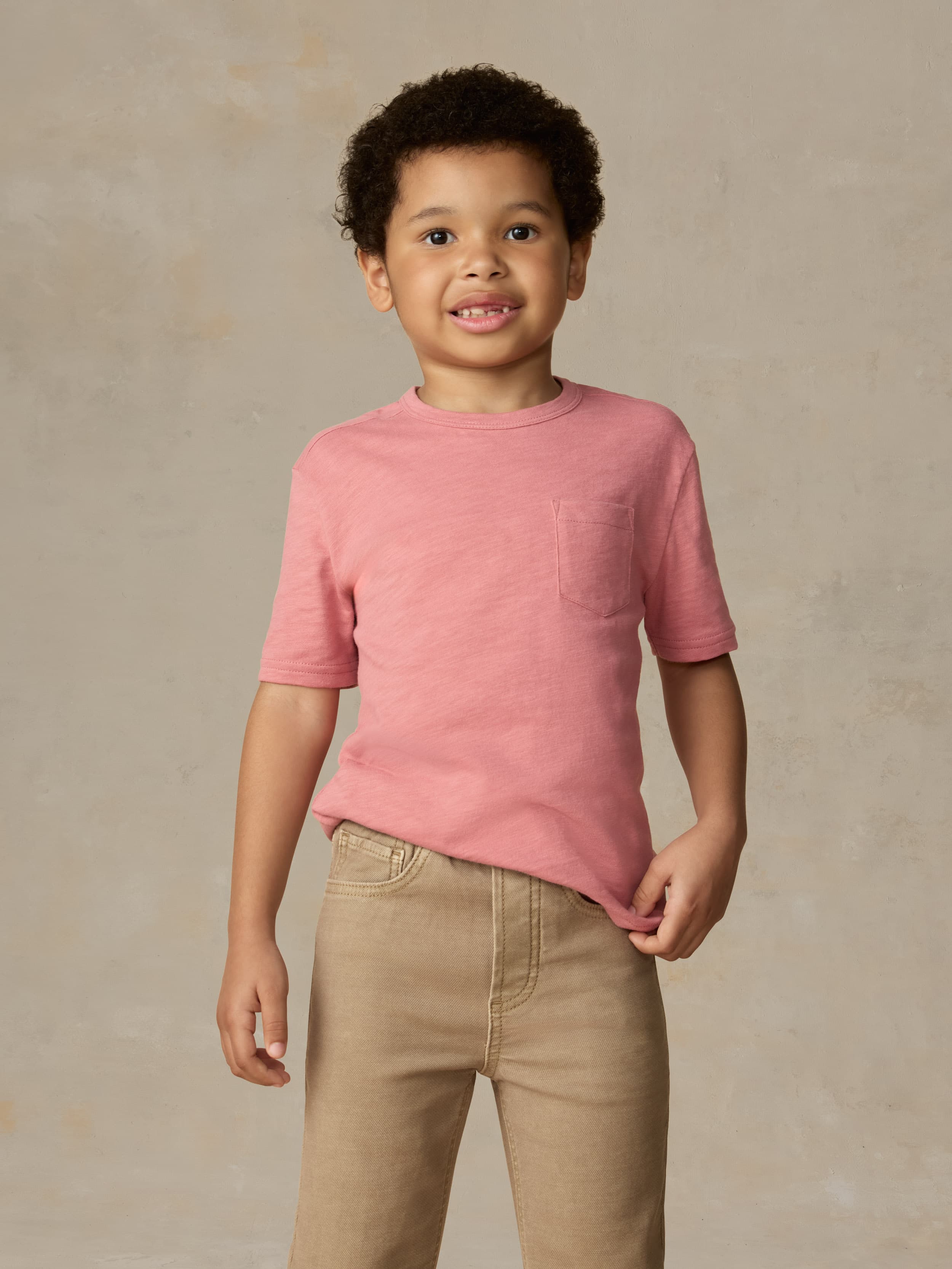 Child wearing a red pocket tee and beige pants against a beige background