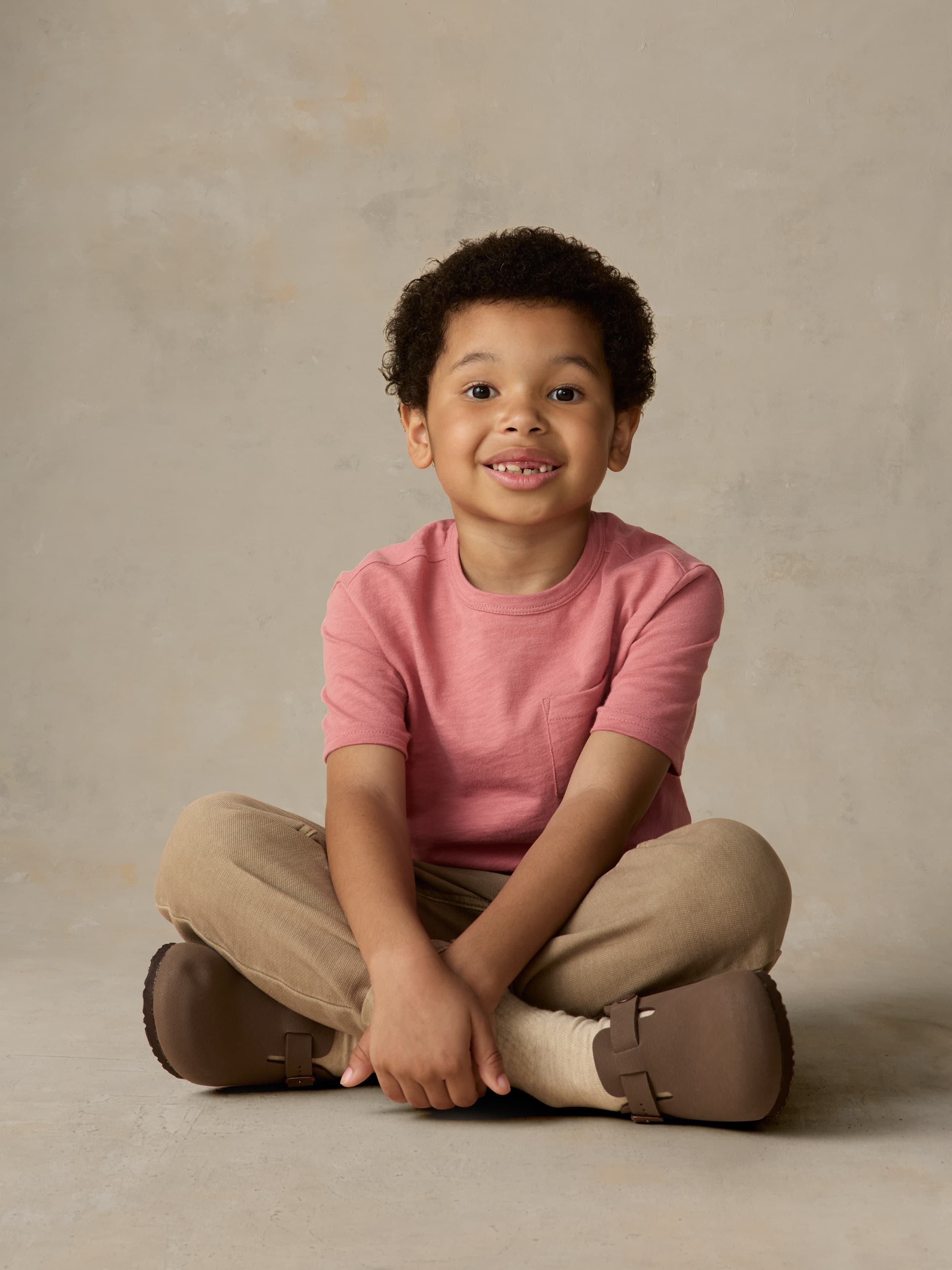 Child wearing a red pocket tee and beige pants against a beige background