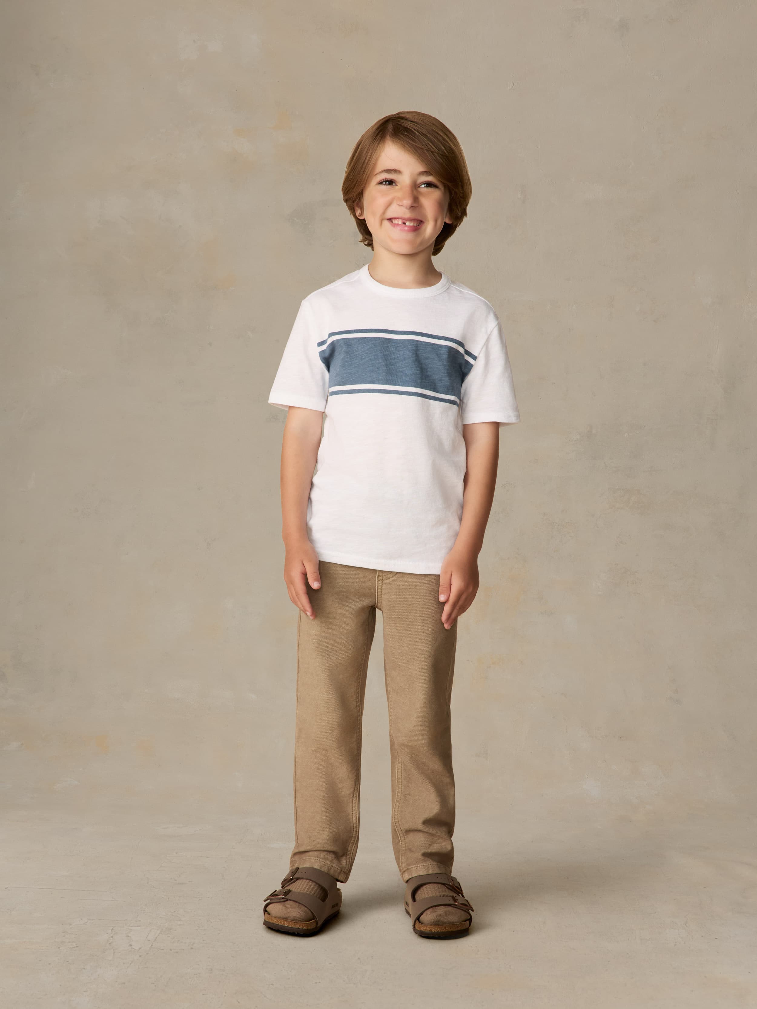 Child wearing a white shirt with blue stripe and beige pants against a beige background