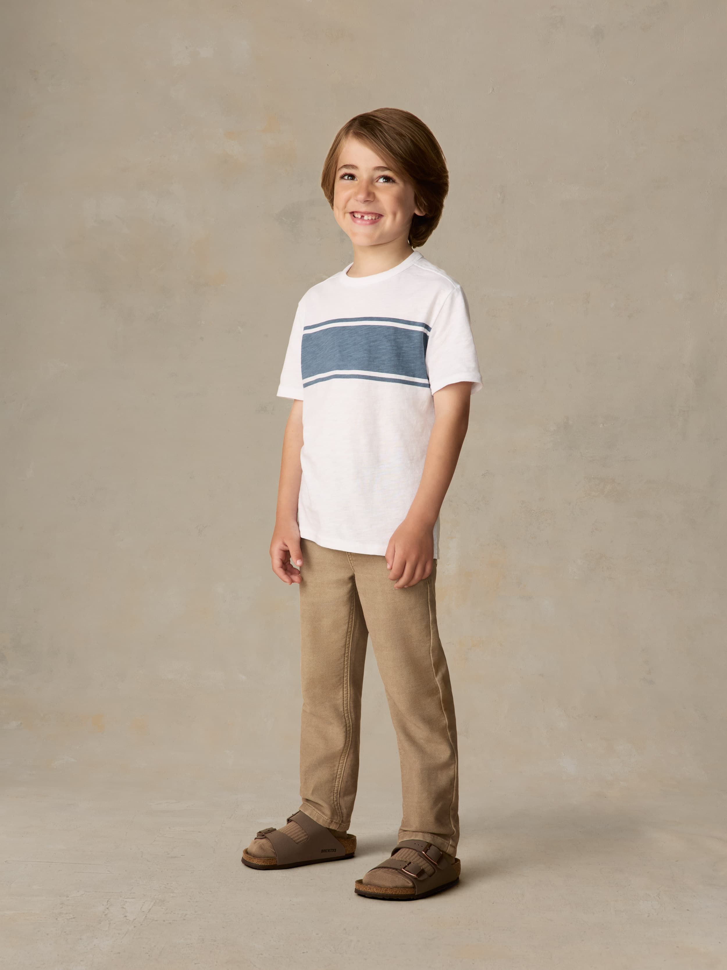 Child wearing a white shirt with blue stripe and beige pants against a beige background