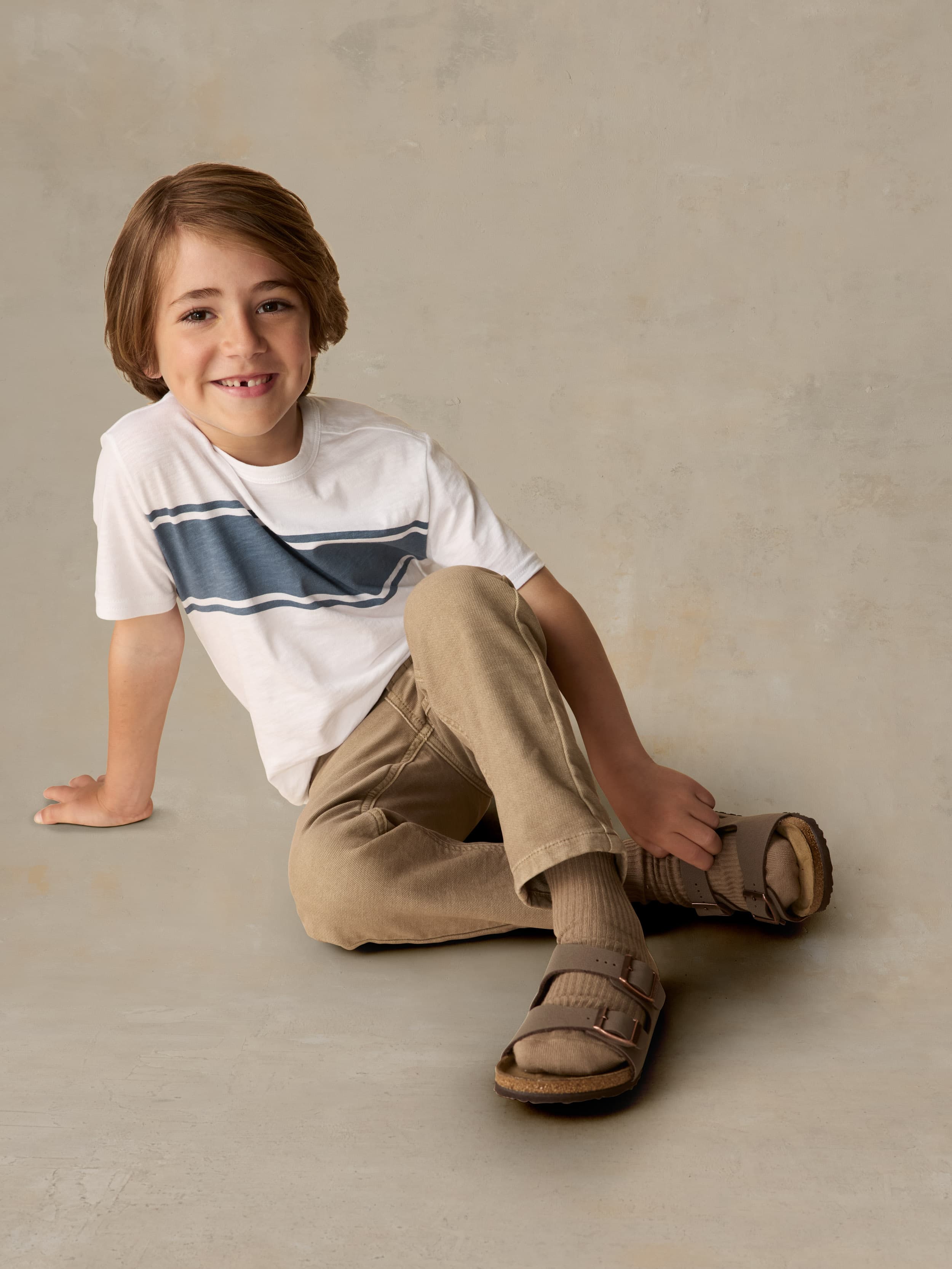 Child wearing a white shirt with blue stripe and beige pants against a beige background