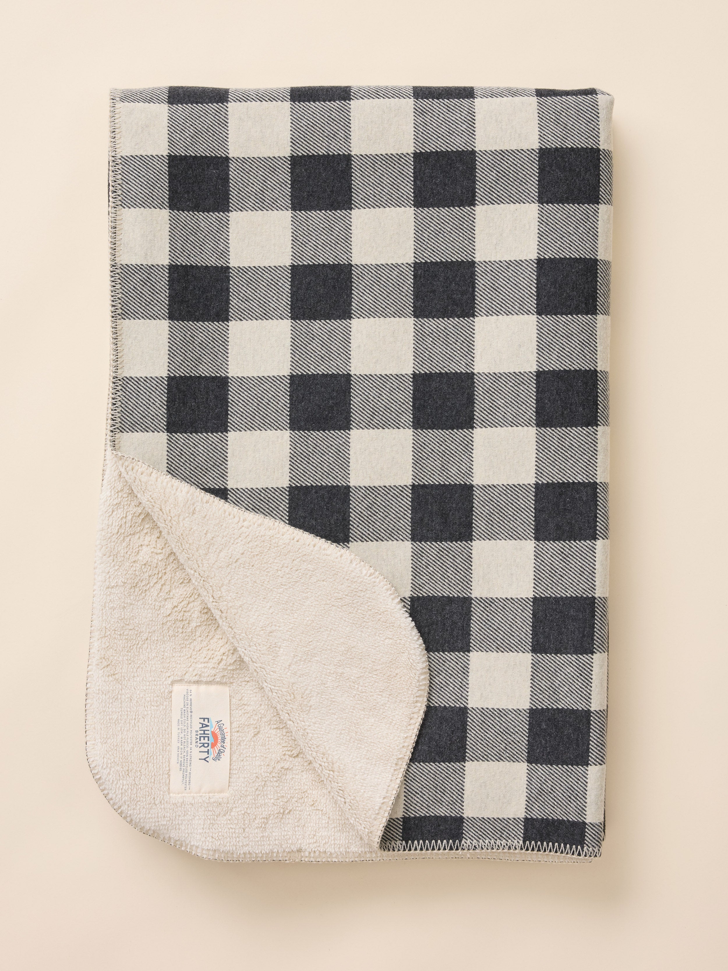 A folded checkered blanket with a black and off-white buffalo plaid pattern, showing a soft off-white fleece lining on the reverse side, photographed on a solid light beige background.