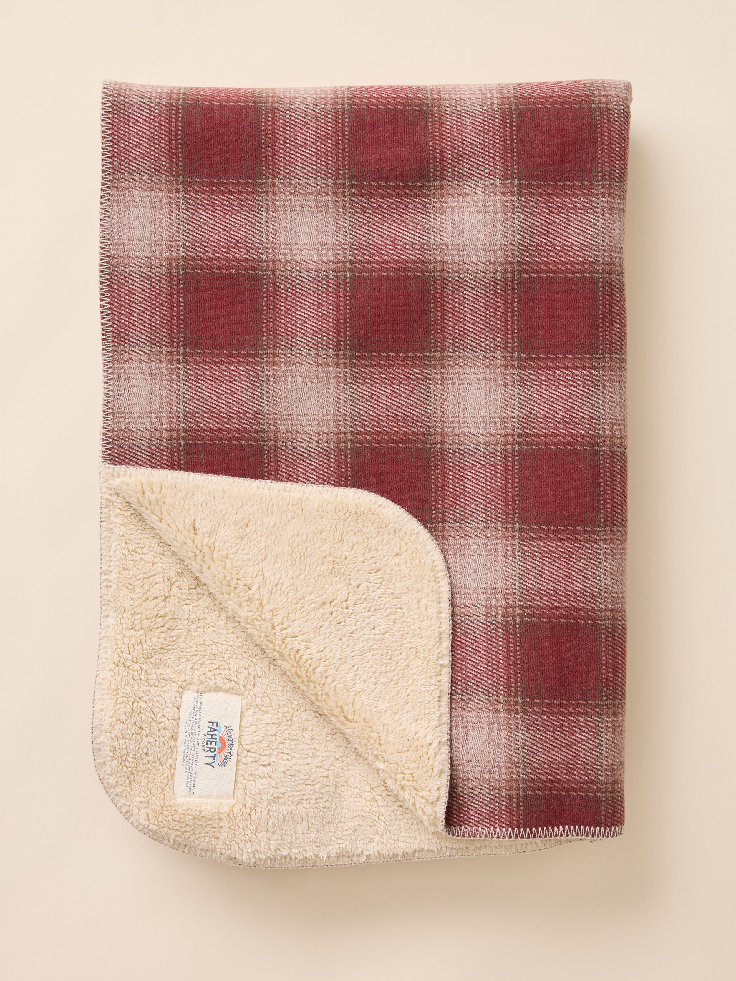 A folded fleece blanket with a red and beige plaid pattern on one side and a soft cream sherpa backing on the other, displayed on a light beige background.