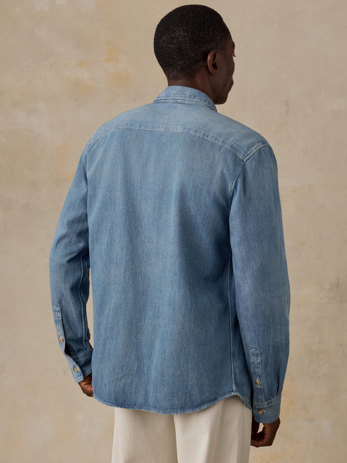 A man viewed from the back wearing a light blue denim long-sleeve button-up shirt and matching blue jeans, standing against a plain beige wall., image 3 of 5