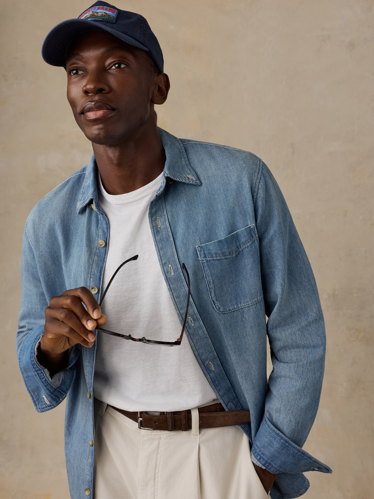 Man wearing a light blue denim shirt over a white t-shirt, khaki pants, and a hat against a beige background, image 4 of 5