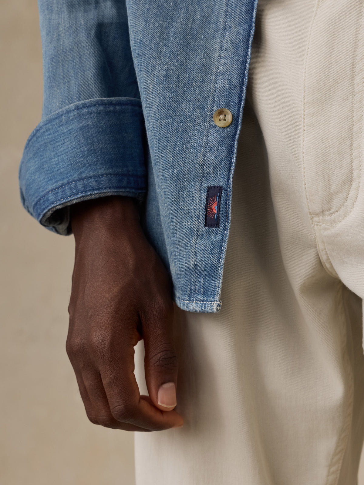 Close-up of a light blue denim button-up shirt showing the button closure, fabric texture, and a small Faherty brand sun and waves patch near the hem, image 5 of 5