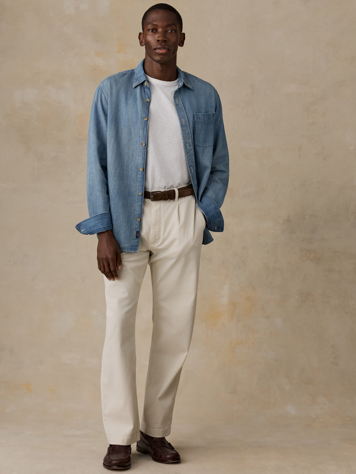 A man wearing a light blue denim shirt and matching light blue jeans, styled with a brown belt and brown loafers, standing against a textured beige wall., image 2 of 5