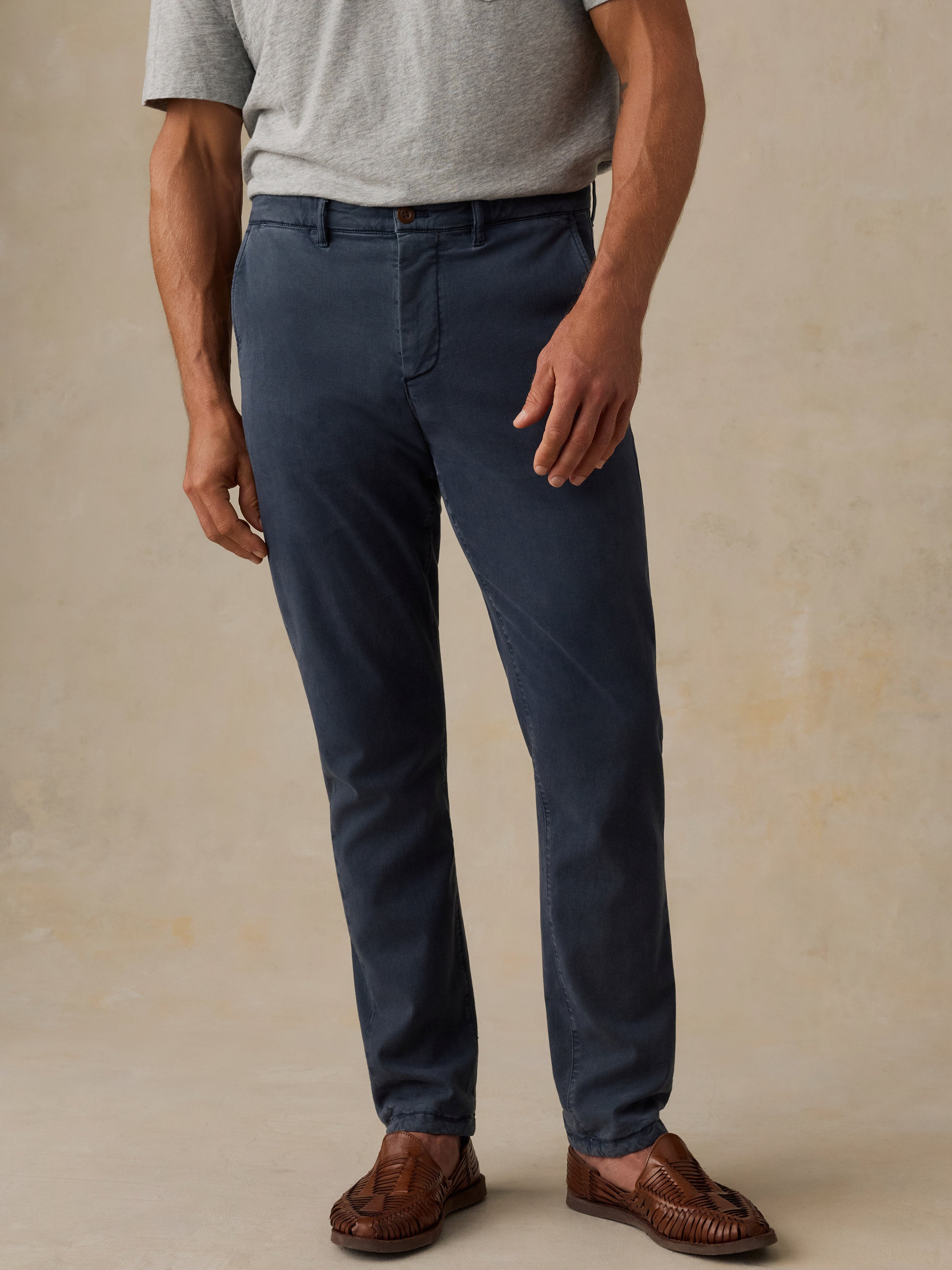 A man wearing slim-fit navy blue dress pants, a light gray t-shirt, and brown leather loafers, standing against a textured beige studio backdrop.