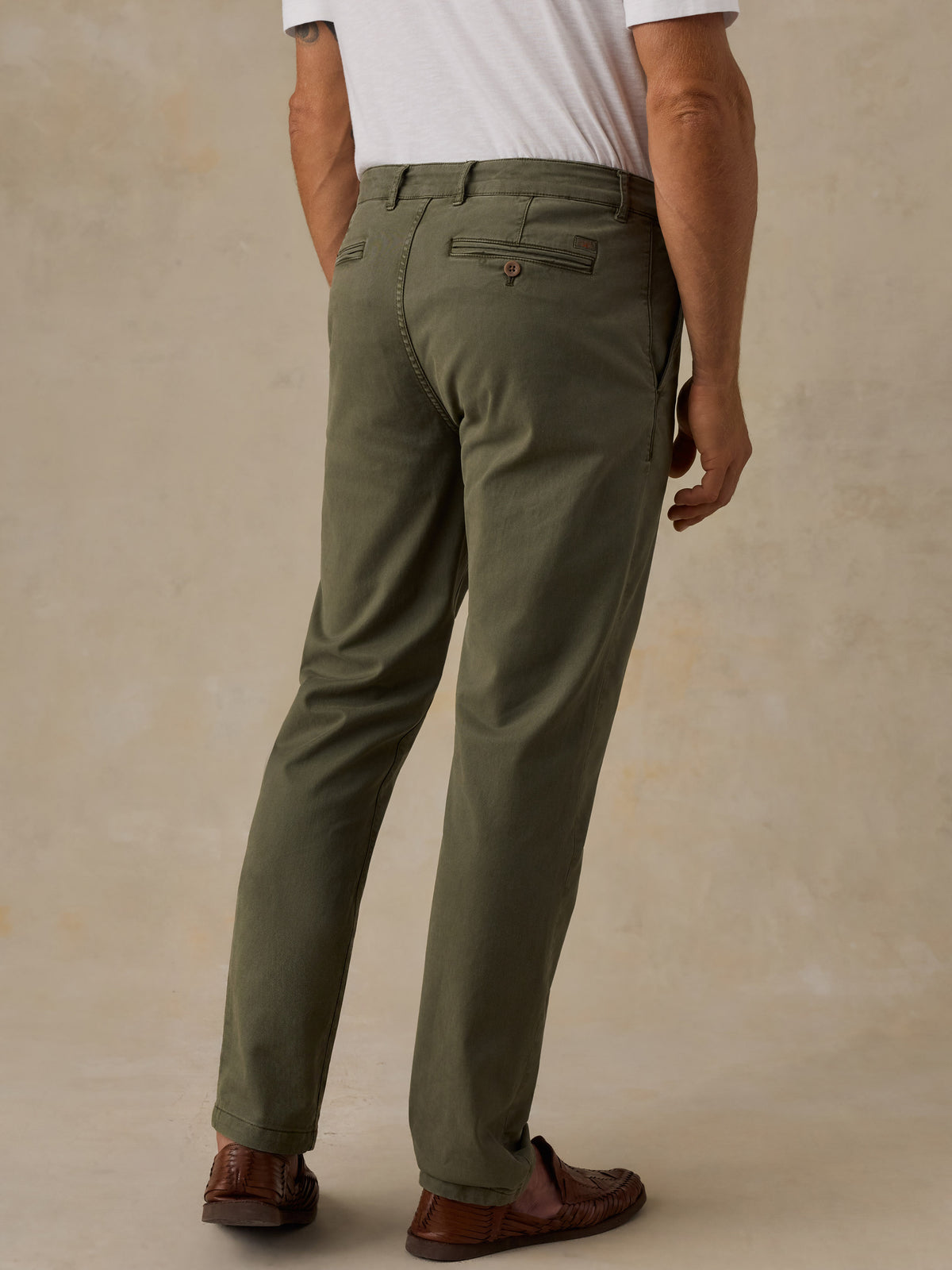 A man wearing olive green chino pants, a white t-shirt, and brown leather loafers, shown from a rear three-quarter angle against a textured beige studio backdrop., image 3 of 5
