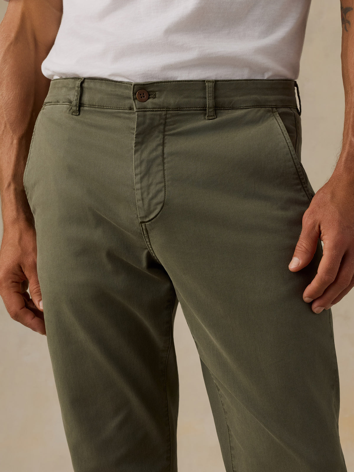 A close-up of a man wearing olive green chinos with a white t-shirt, showing the front waist, belt loops, button, and front pockets., image 5 of 5