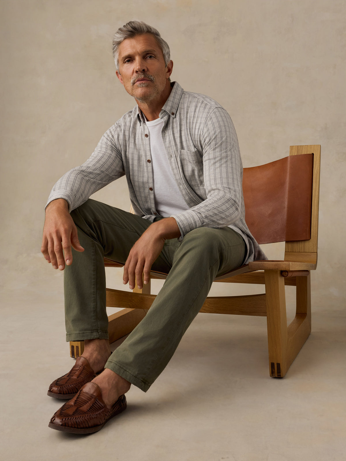 A man sits on a wooden chair with brown leather backrests, wearing a light gray and white striped button-down shirt over a white undershirt, olive green pants, and brown leather loafers, against a textured beige studio backdrop., image 4 of 5
