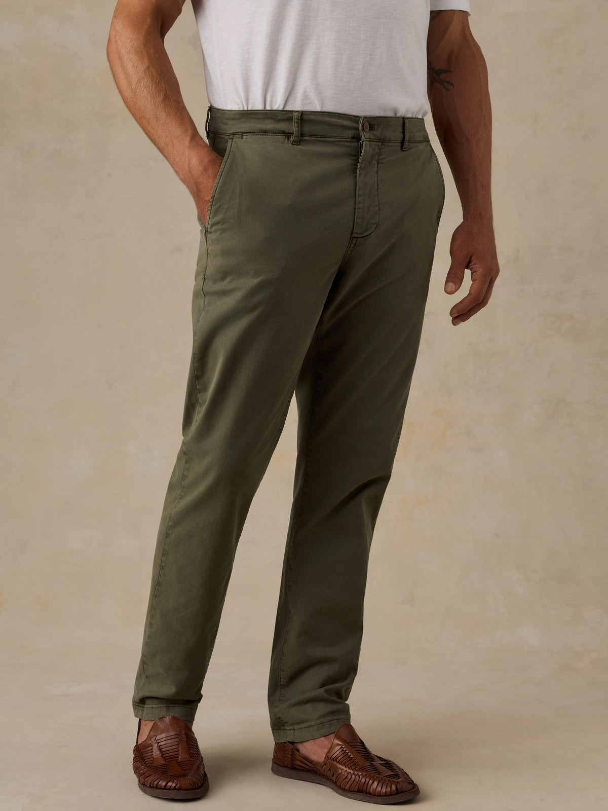 A man wearing olive green slim-fit trousers, a white t-shirt, and brown leather loafers, standing against a textured beige studio background., image 1 of 5