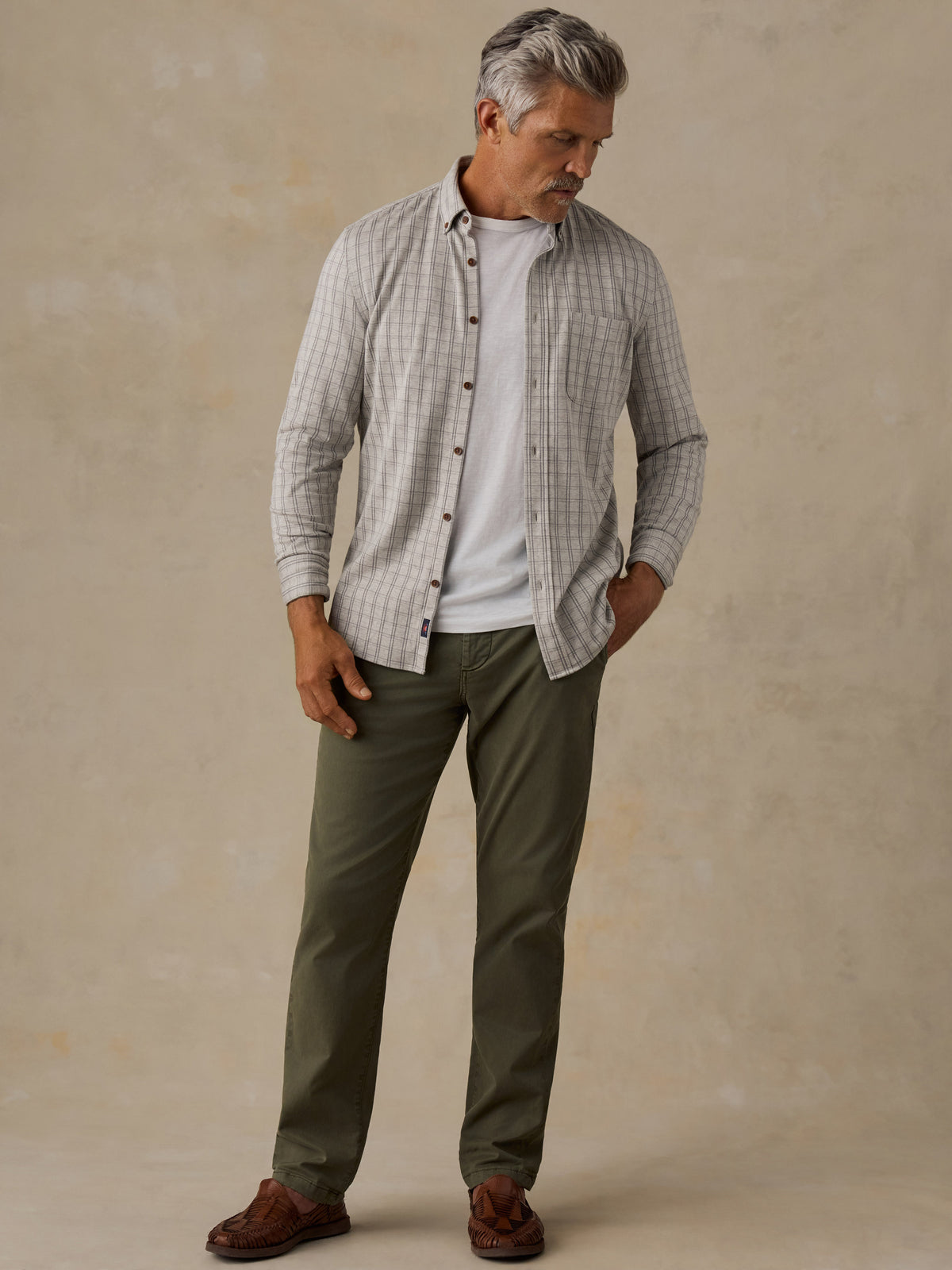 A man wearing a light gray and white checkered button-down shirt over a white t-shirt, paired with olive green pants and brown shoes, standing against a textured beige studio backdrop., image 2 of 5