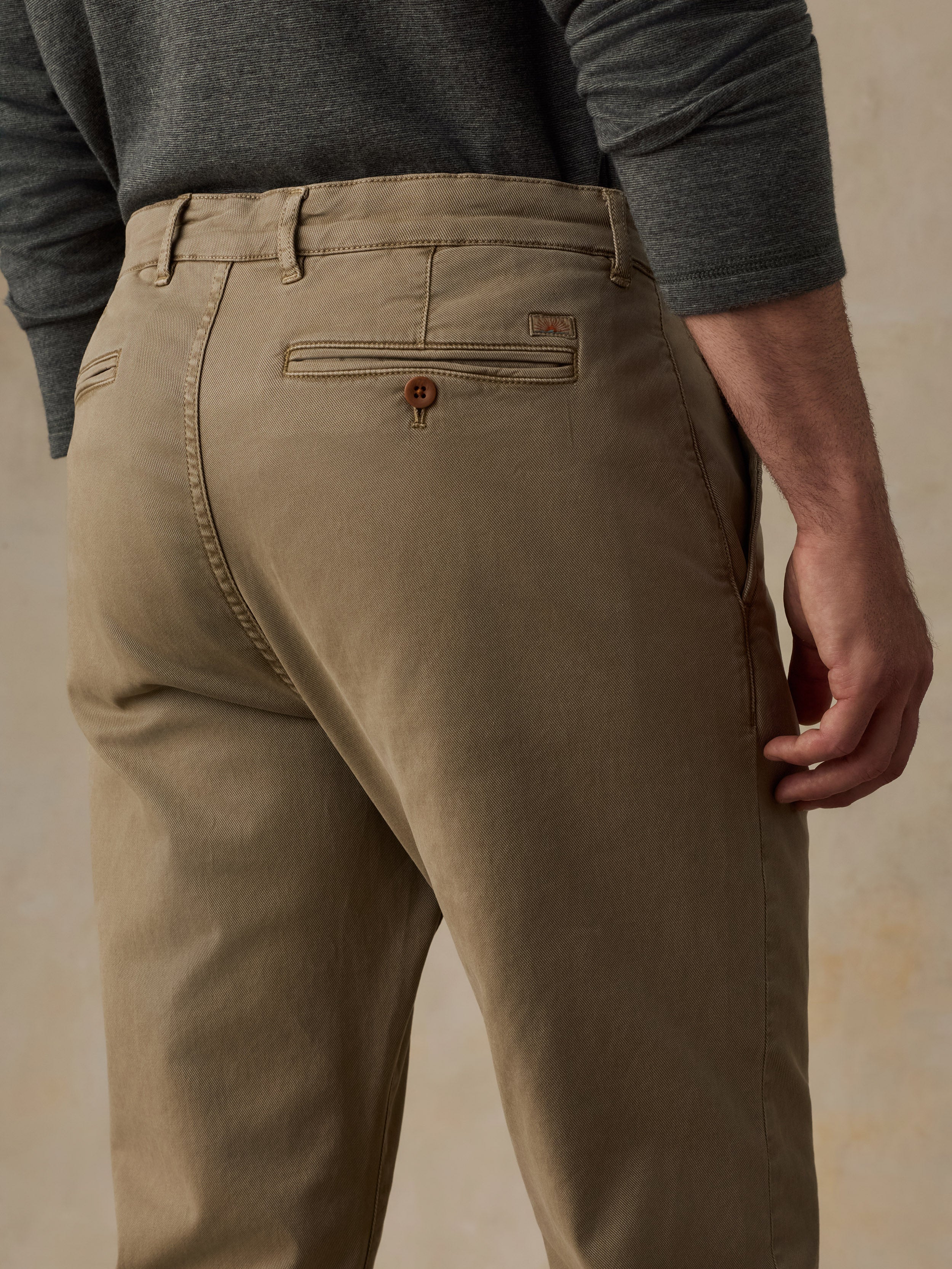 Close-up back view of a man wearing tan chinos with a dark gray long-sleeve shirt, showing the back pocket and waistband details against a neutral beige background.