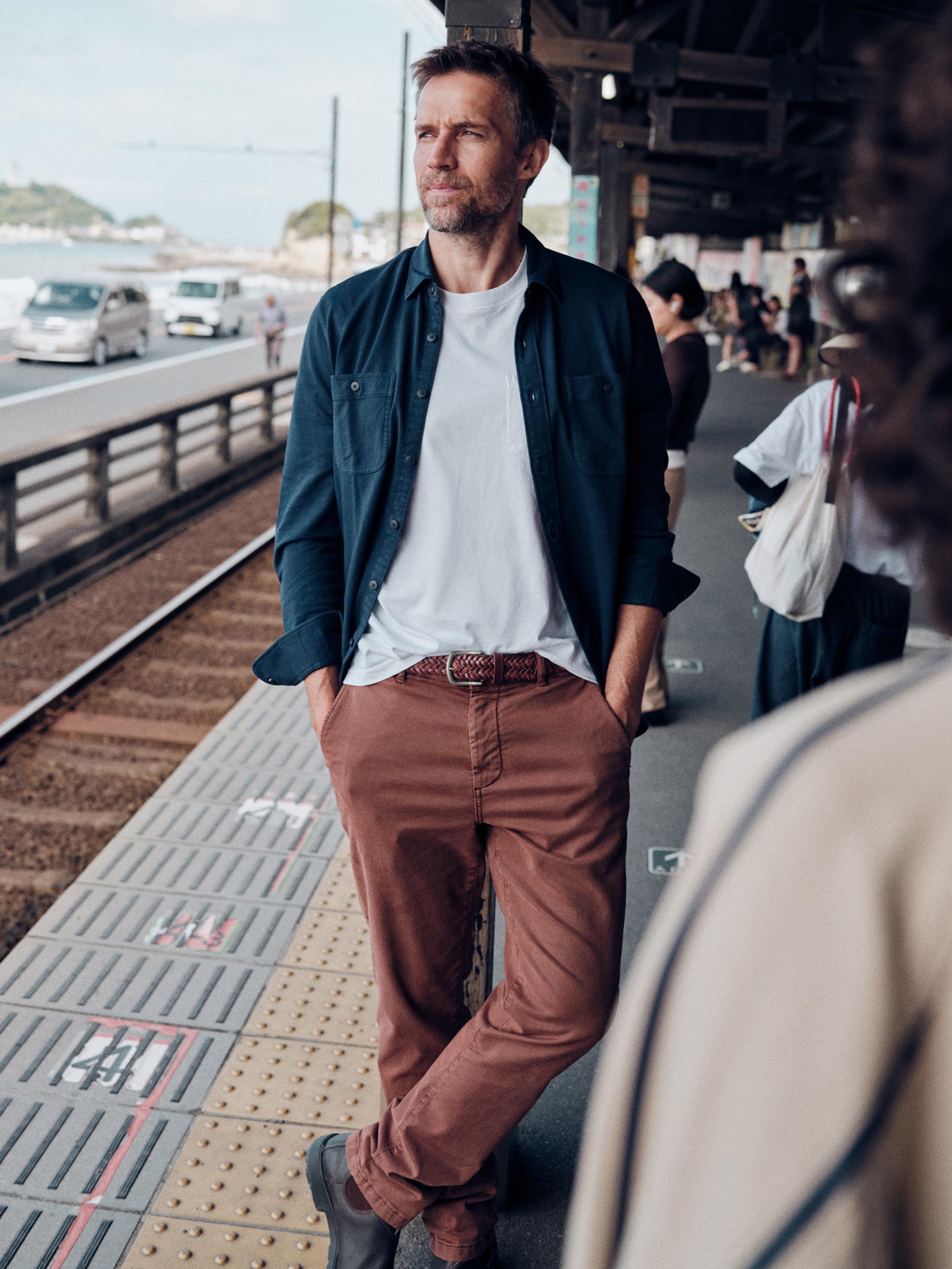 A man standing on a train station platform wearing a dark navy overshirt over a white t-shirt, brown trousers, and dark shoes. The background shows a blurred train station environment with tracks, cars, and other passengers.
