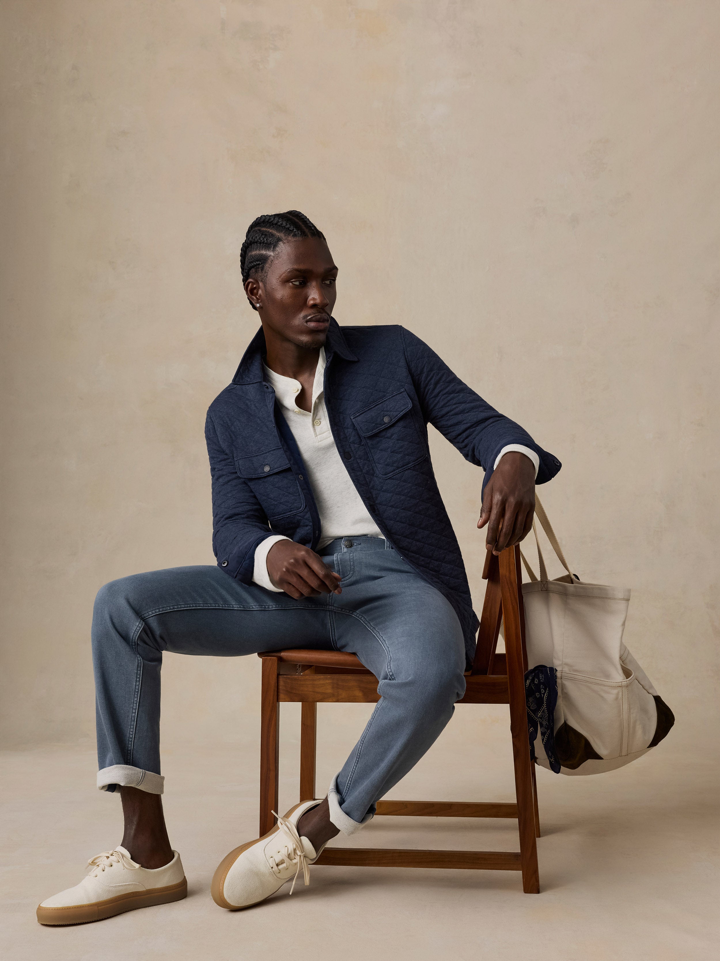 A man sits on a wooden chair wearing a navy quilted jacket, a white polo shirt, blue jeans, and cream-colored sneakers.