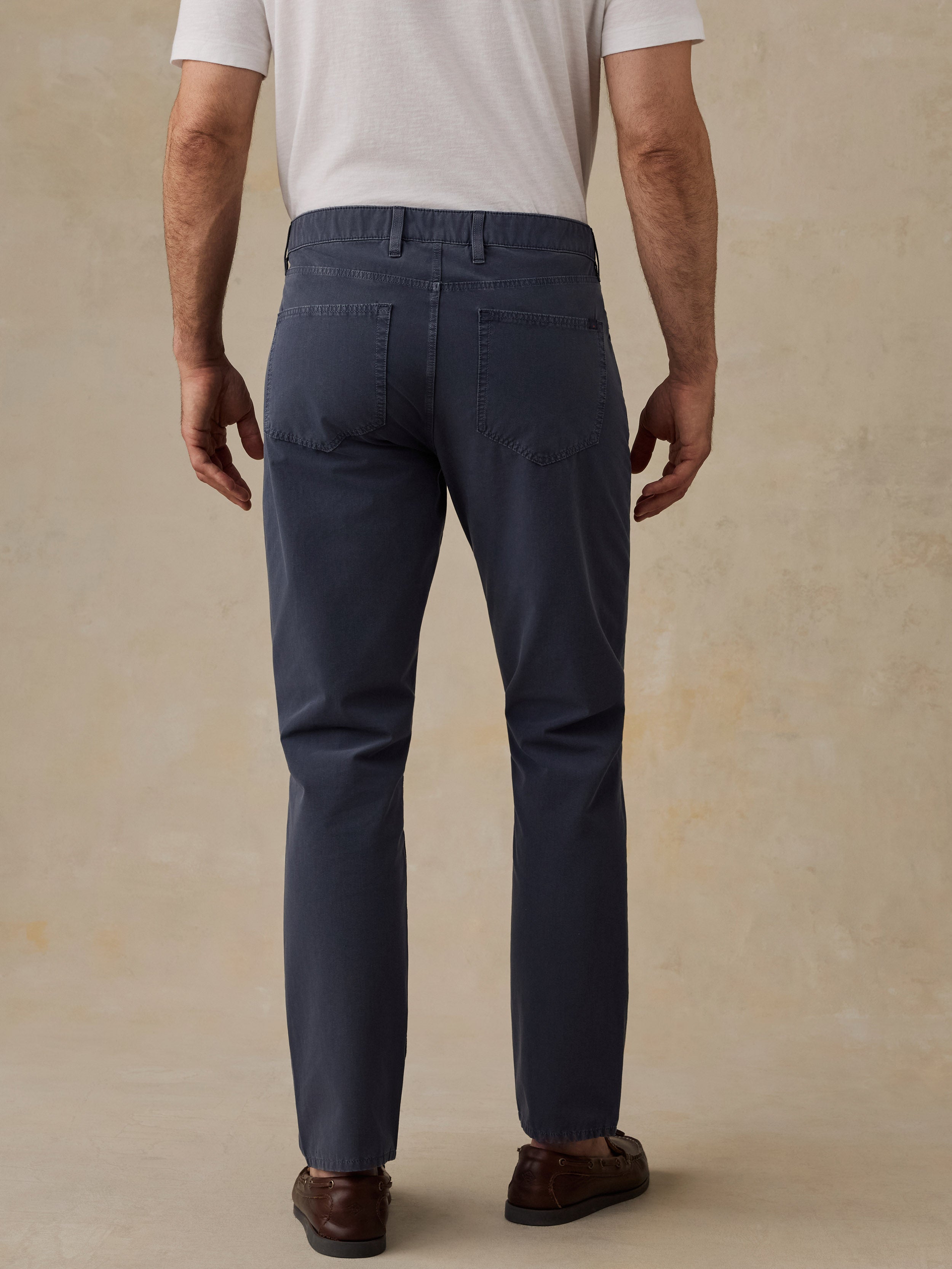 Rear view of a man wearing navy blue slim-fit trousers, a white t-shirt, and brown leather loafers, standing against a textured beige studio backdrop.