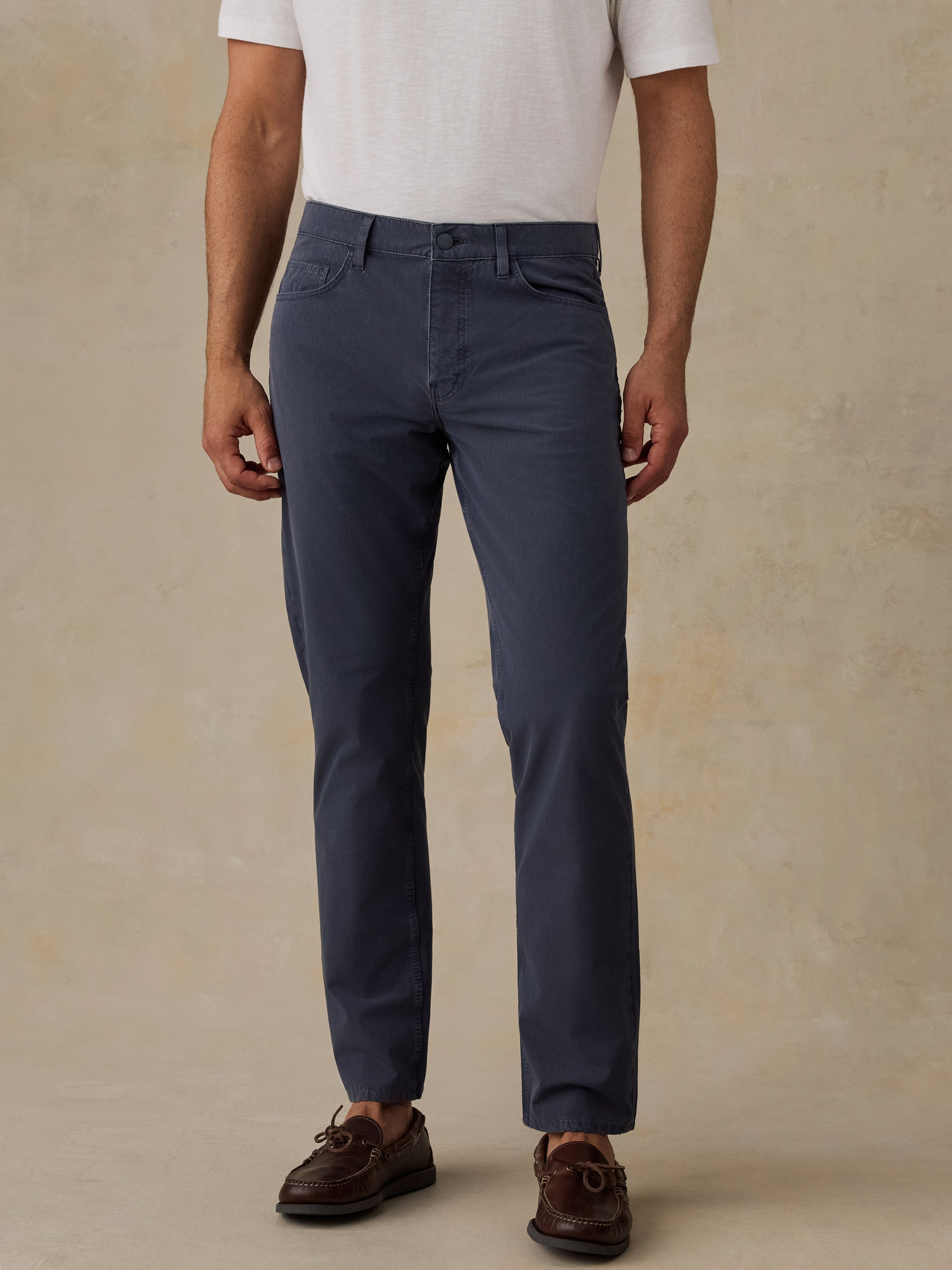 A man wearing slim-fit navy blue trousers, a white t-shirt, and brown leather boat shoes, standing against a textured beige studio backdrop.