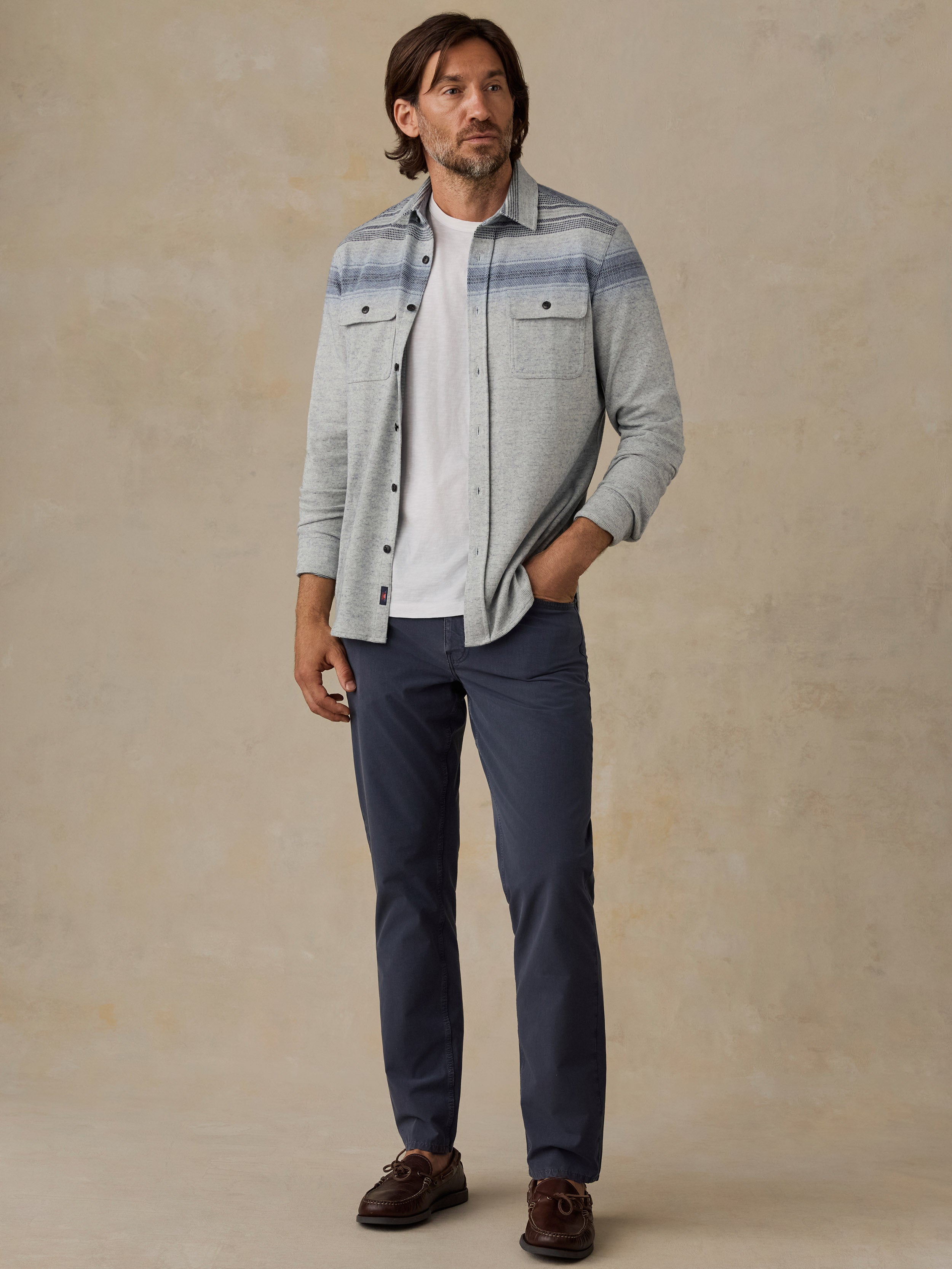 A man wearing a light gray heathered button-down shirt with blue stripes, a white t-shirt, navy blue pants, and brown leather shoes, standing against a textured beige background.