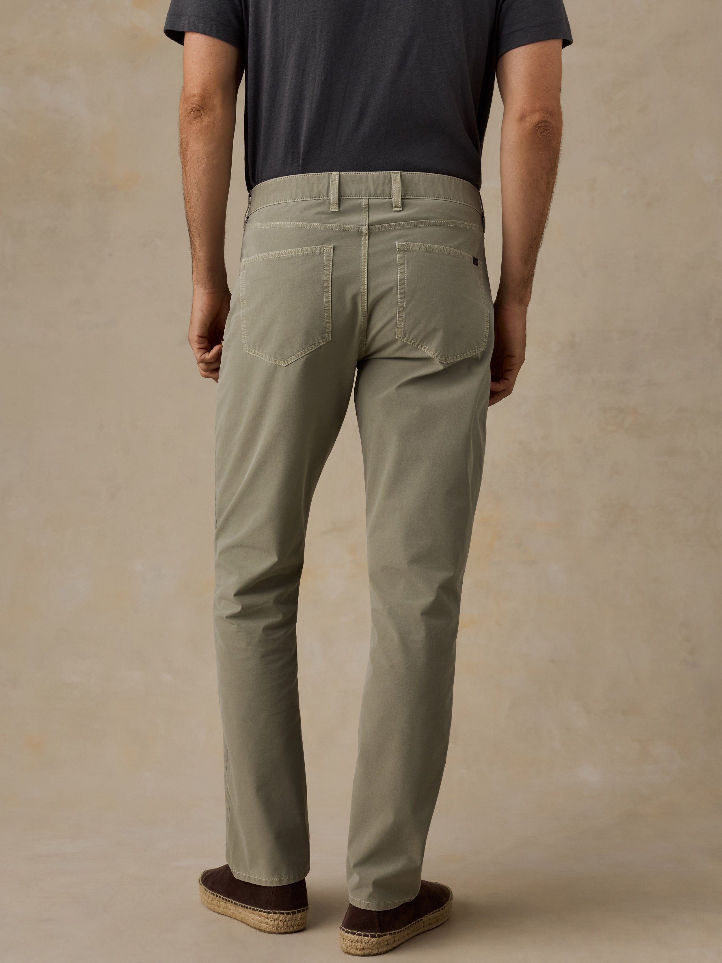 A man viewed from the back wearing slim-fit khaki trousers, a dark gray t-shirt, and brown espadrille shoes, standing against a textured beige studio backdrop.