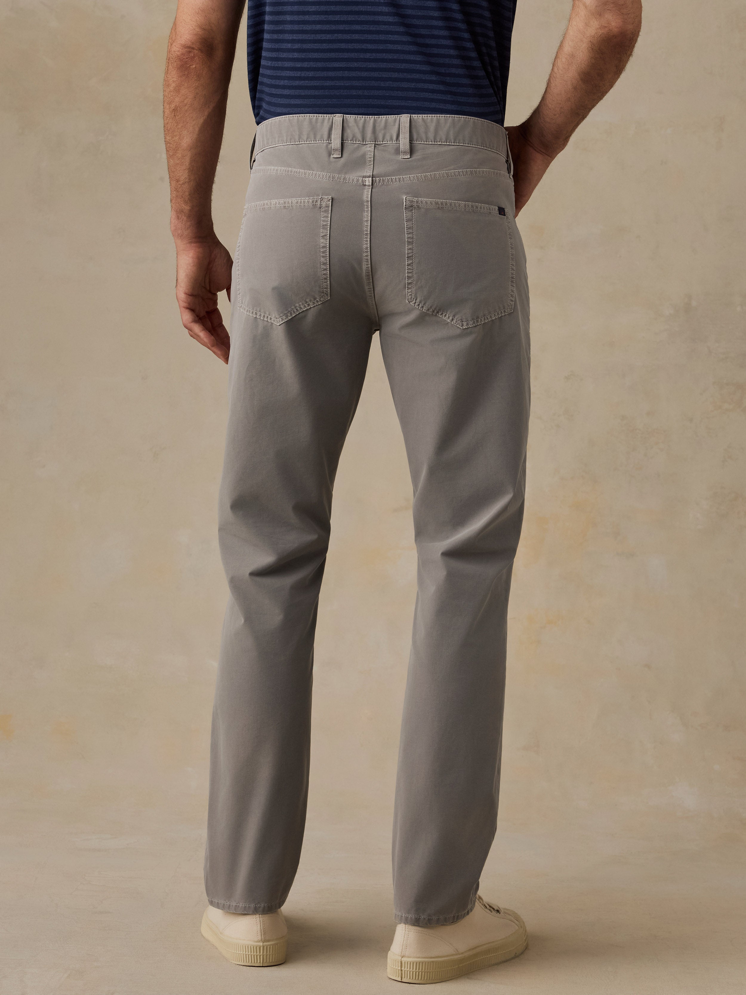 A man viewed from the back wearing light gray slim-fit trousers, a navy blue striped t-shirt, and cream-colored sneakers, standing against a textured beige studio backdrop.