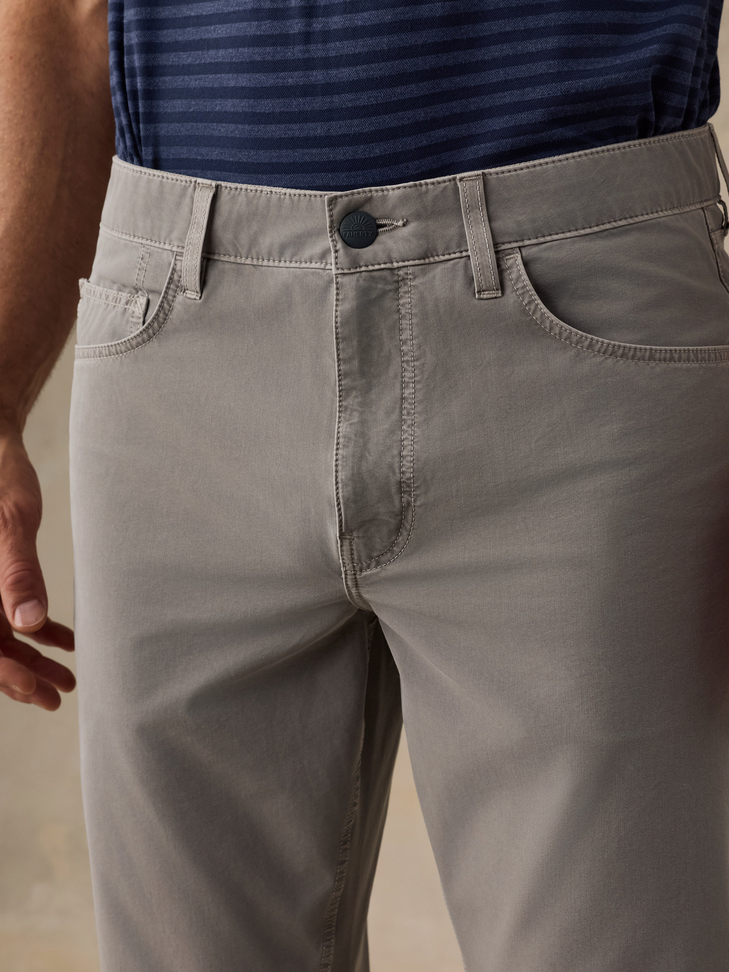 Close-up of a man wearing light gray chino pants, showing the front waistband, button, belt loops, front pockets, and upper thigh area. A navy striped shirt is partially visible at the top.