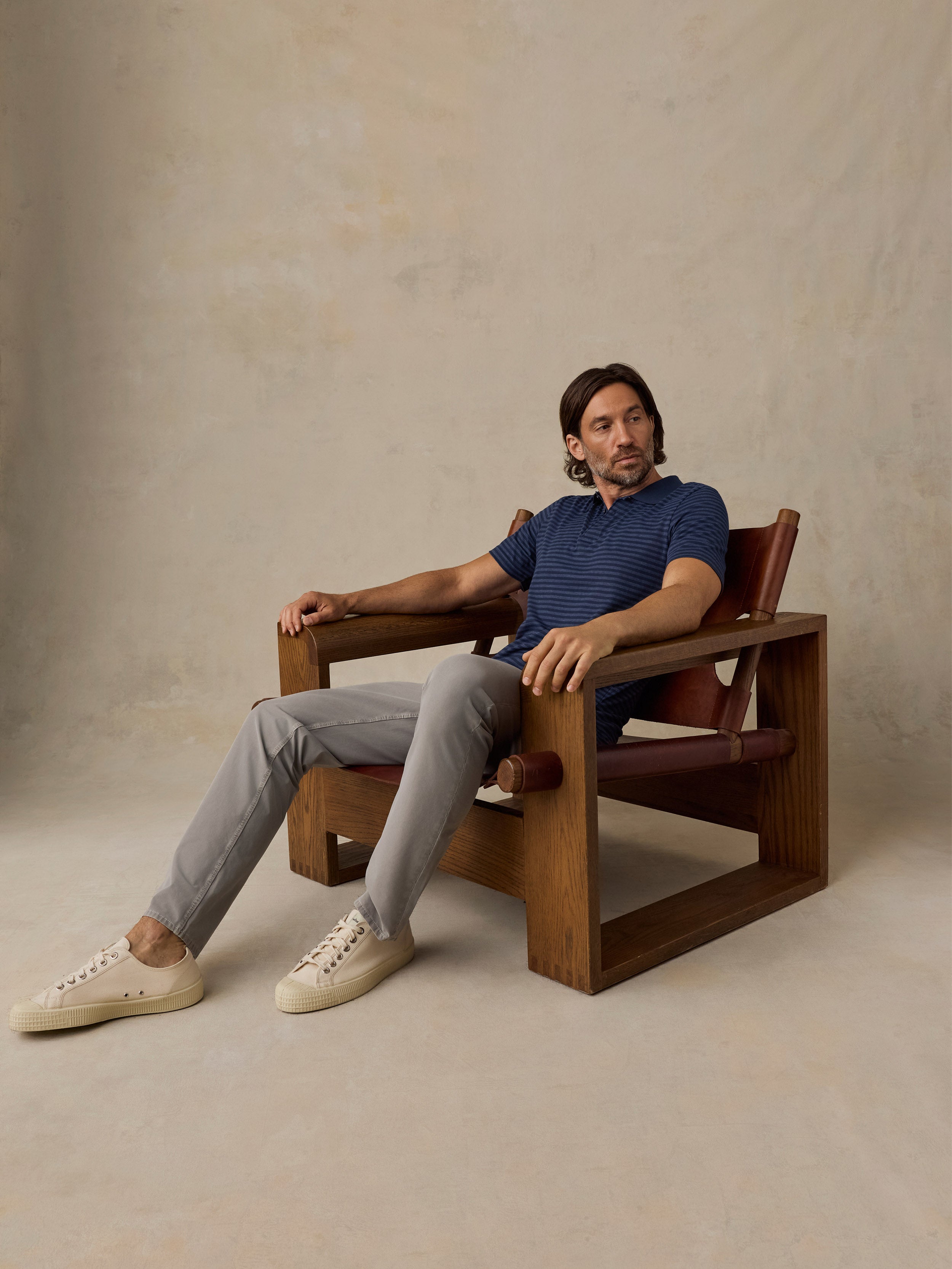 A man sits in a wooden armchair with reddish-brown leather cushions, wearing a blue striped polo shirt, light gray trousers, and cream sneakers, against a textured beige studio backdrop.