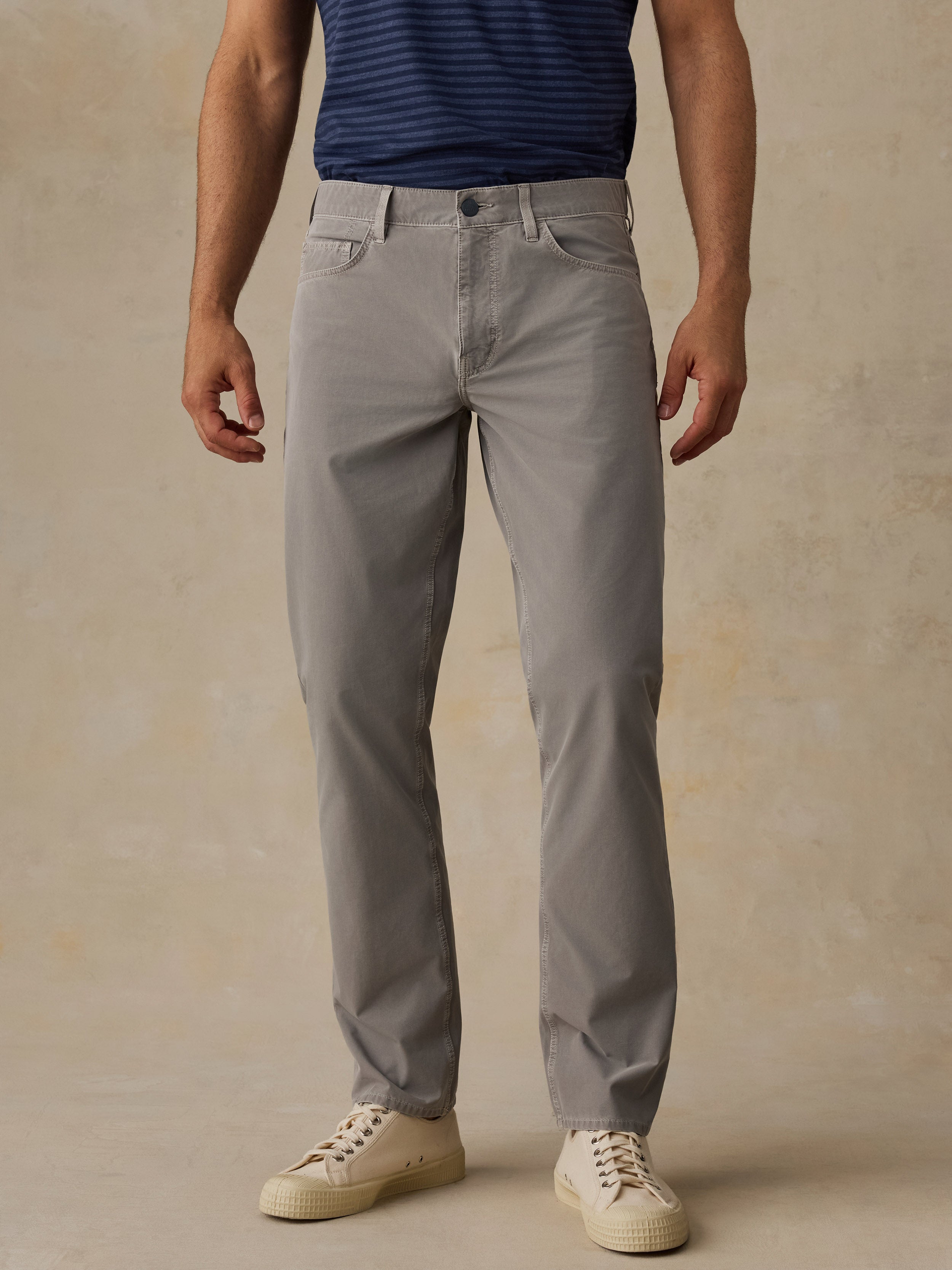 A man wearing light gray slim-fit chino pants, a navy blue striped t-shirt, and cream-colored sneakers, standing against a textured beige studio backdrop.