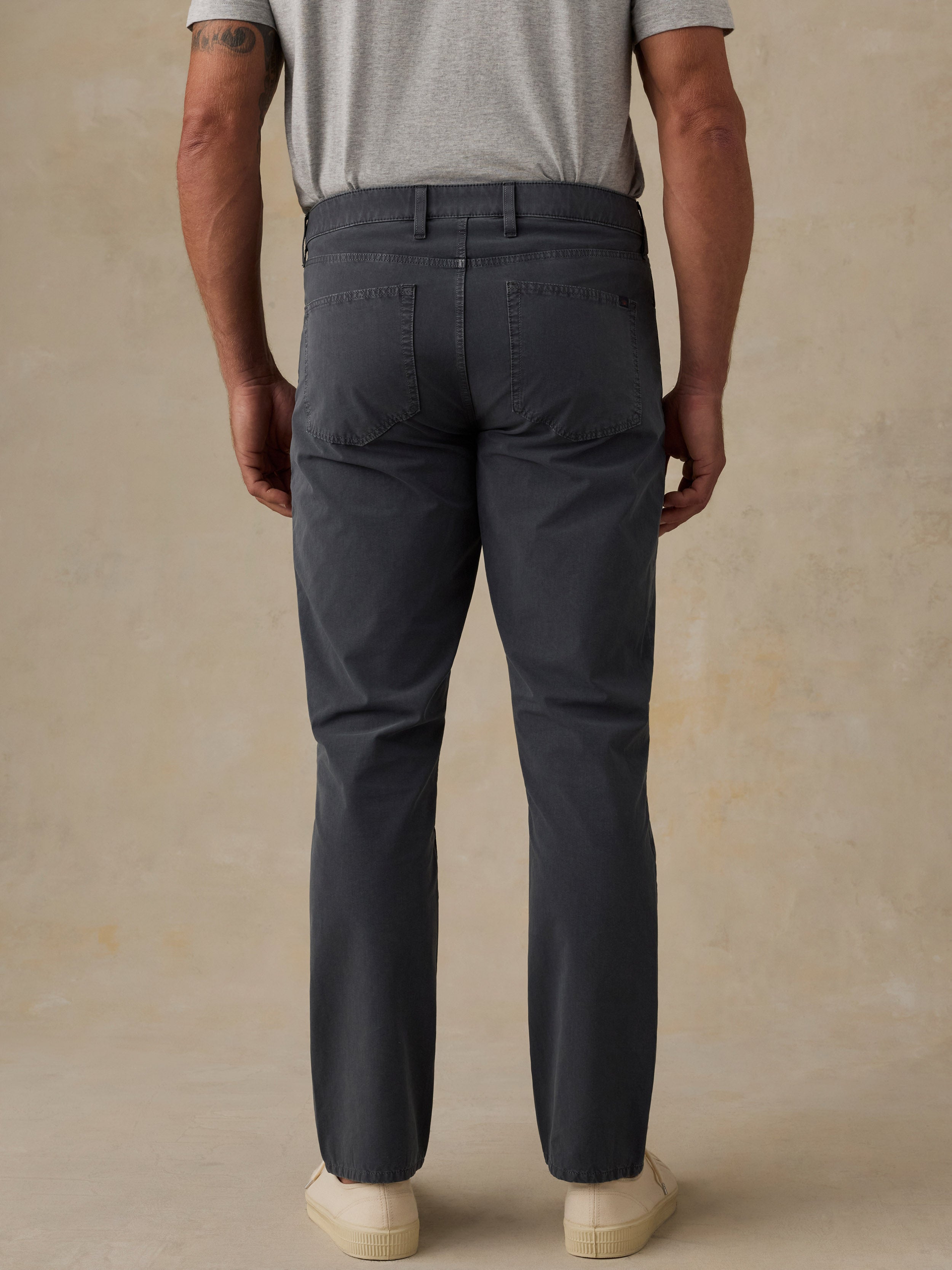 A man viewed from the back wearing dark charcoal gray straight-leg chinos, a light gray t-shirt, and cream sneakers, standing against a textured beige studio backdrop.