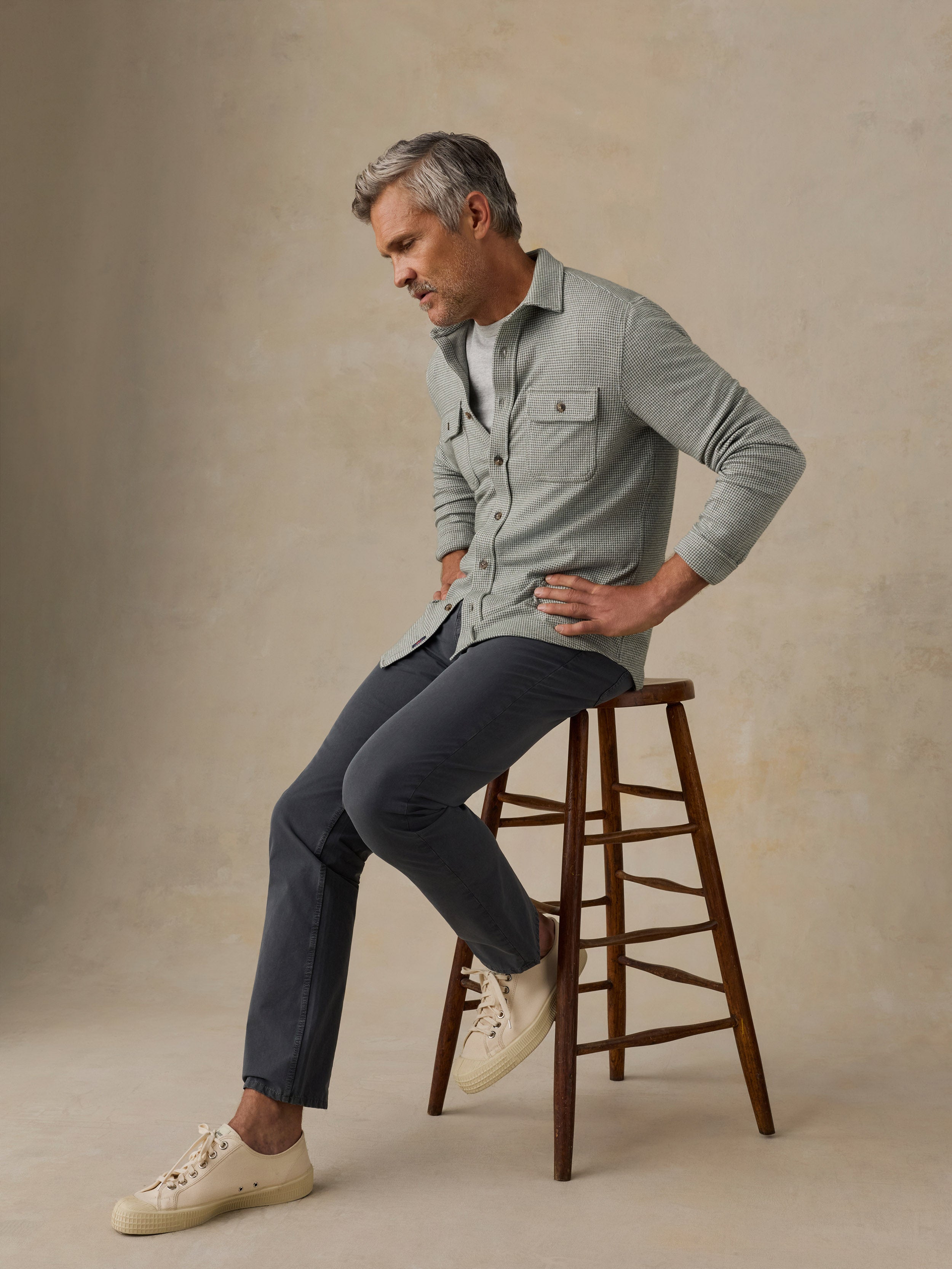 A man with gray hair sits on a wooden stool, wearing a light sage green long-sleeve button-up shirt, dark charcoal trousers, and off-white sneakers, against a textured beige studio backdrop.