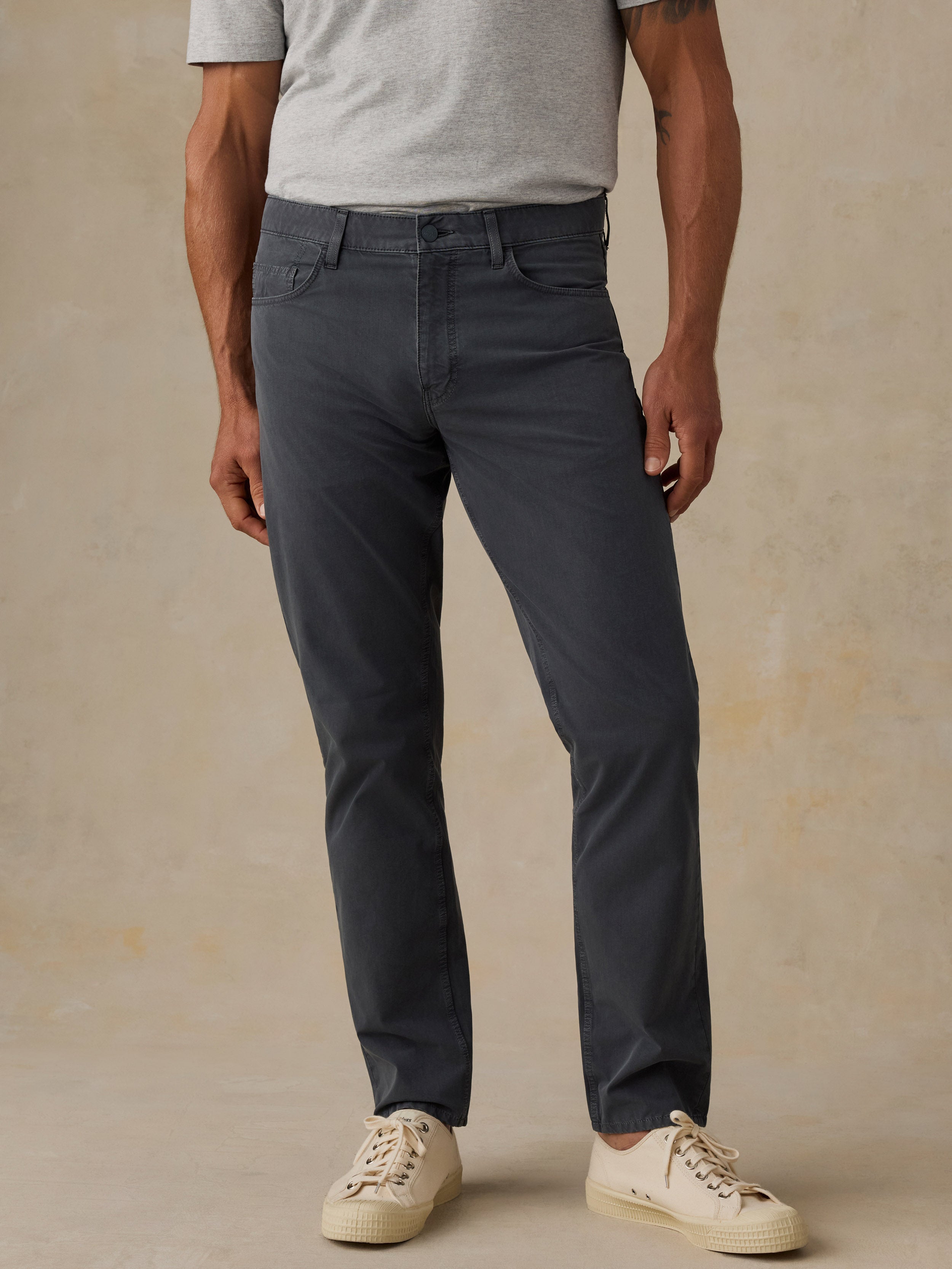 A person wearing slim-fit dark gray pants, a light gray heather t-shirt, and off-white sneakers, standing against a textured beige studio backdrop.