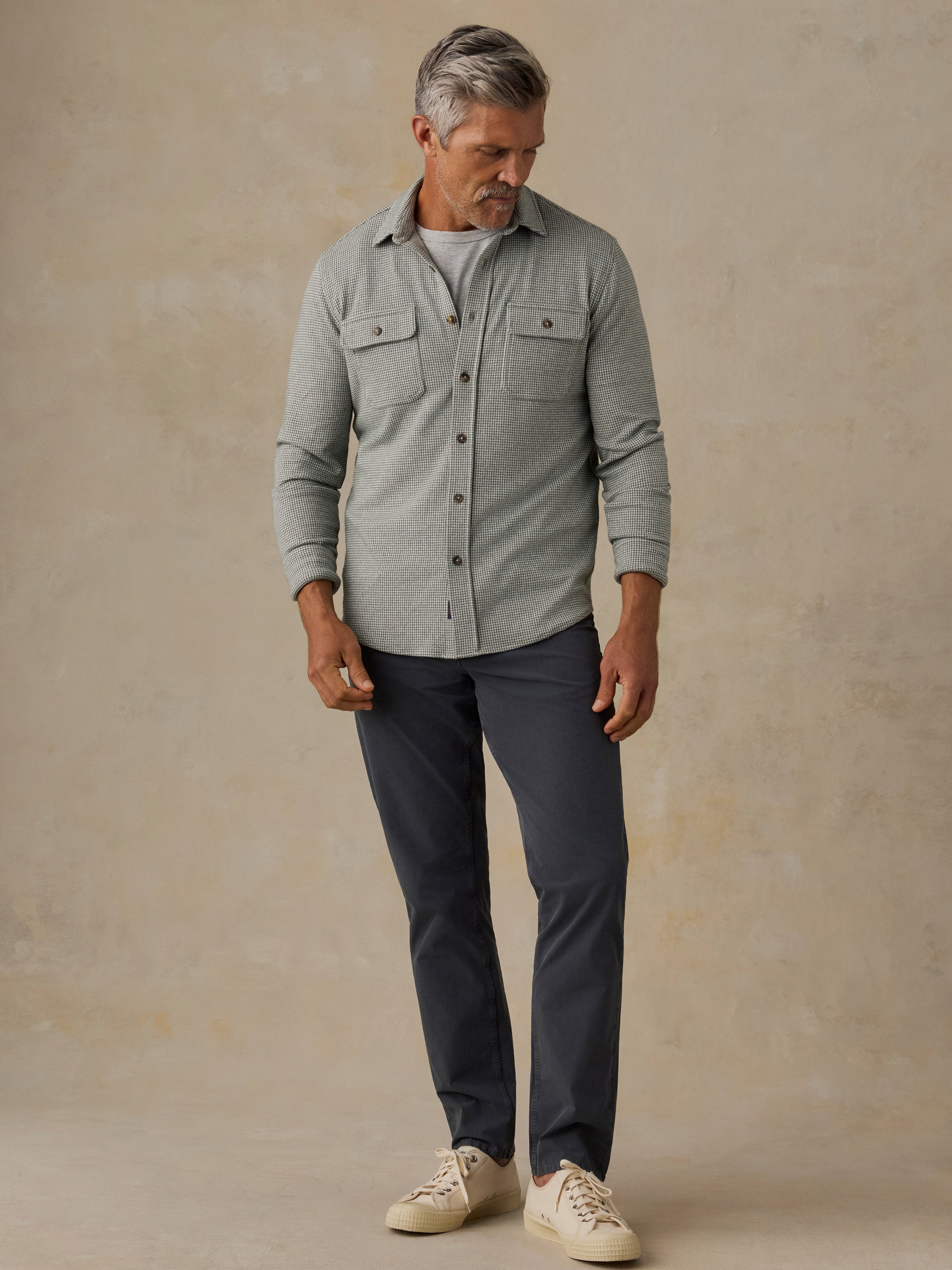 A man wearing a light gray long-sleeve button-up shirt, dark navy pants, and cream sneakers, standing against a textured beige studio backdrop.