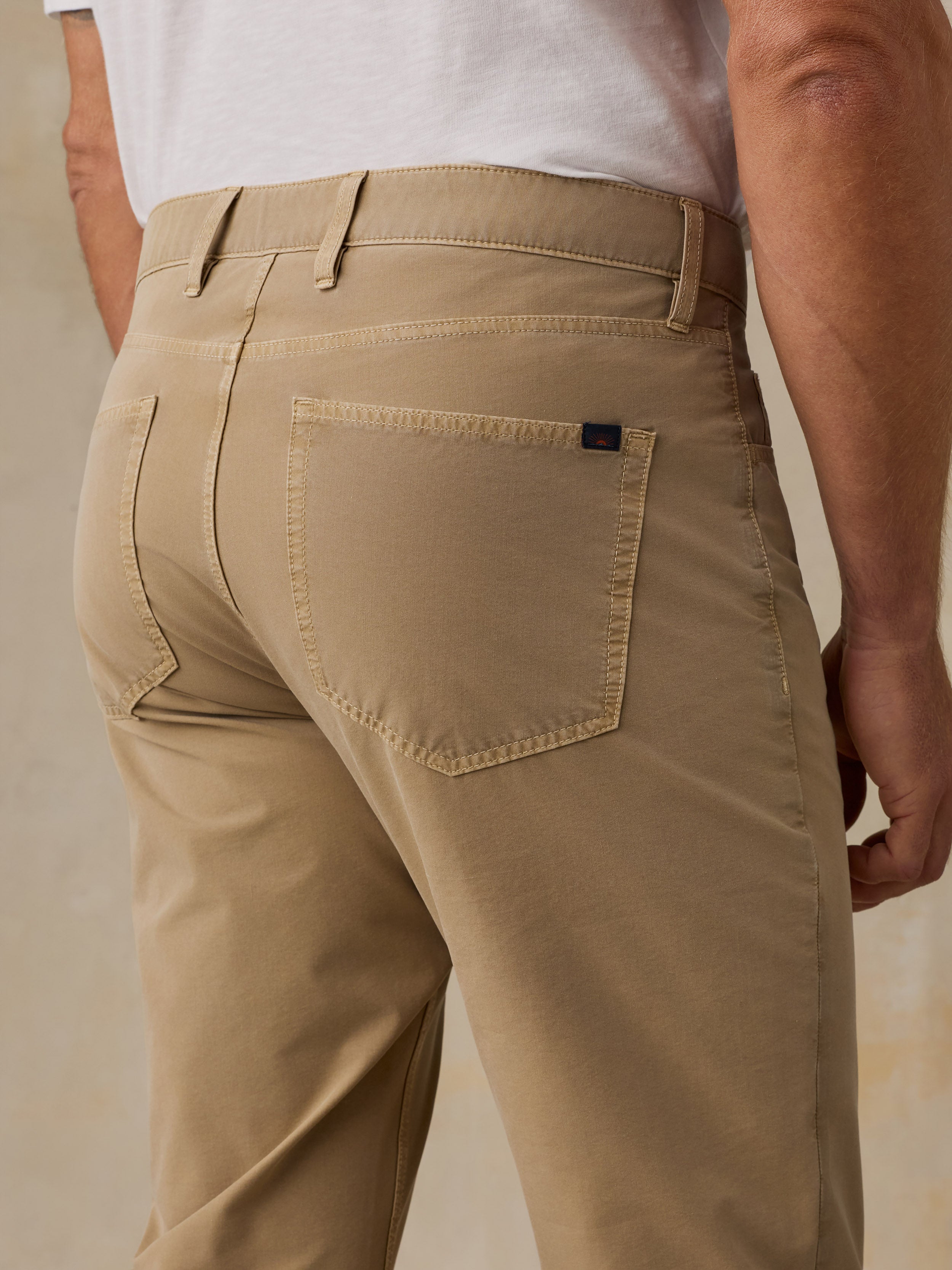 Close-up back view of a man wearing tan chinos or casual trousers with a white shirt partially visible, against a plain beige studio background.