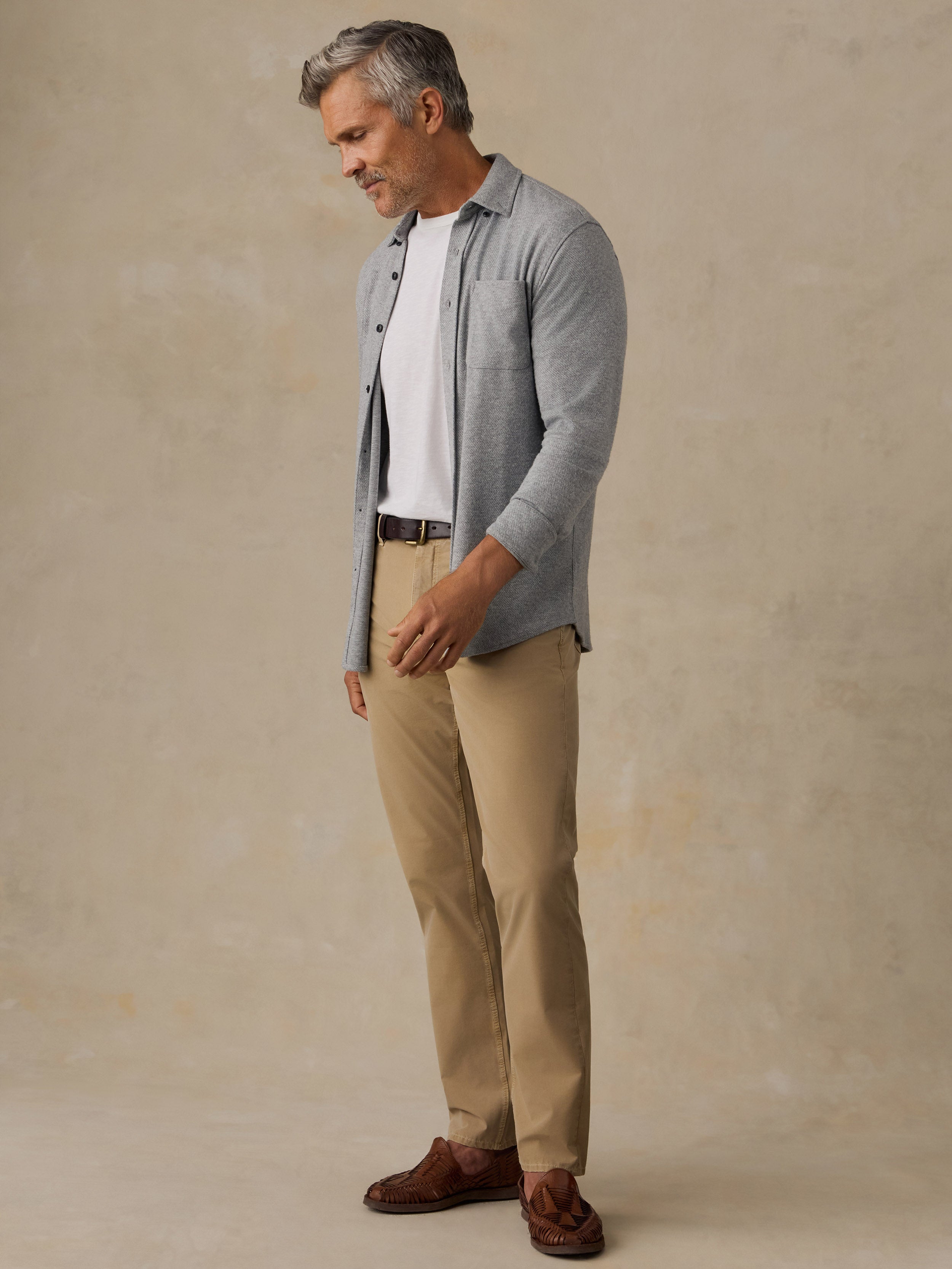 A man wearing a light gray button-down shirt over a white t-shirt, paired with beige trousers and brown suede loafers, standing against a textured beige studio backdrop.