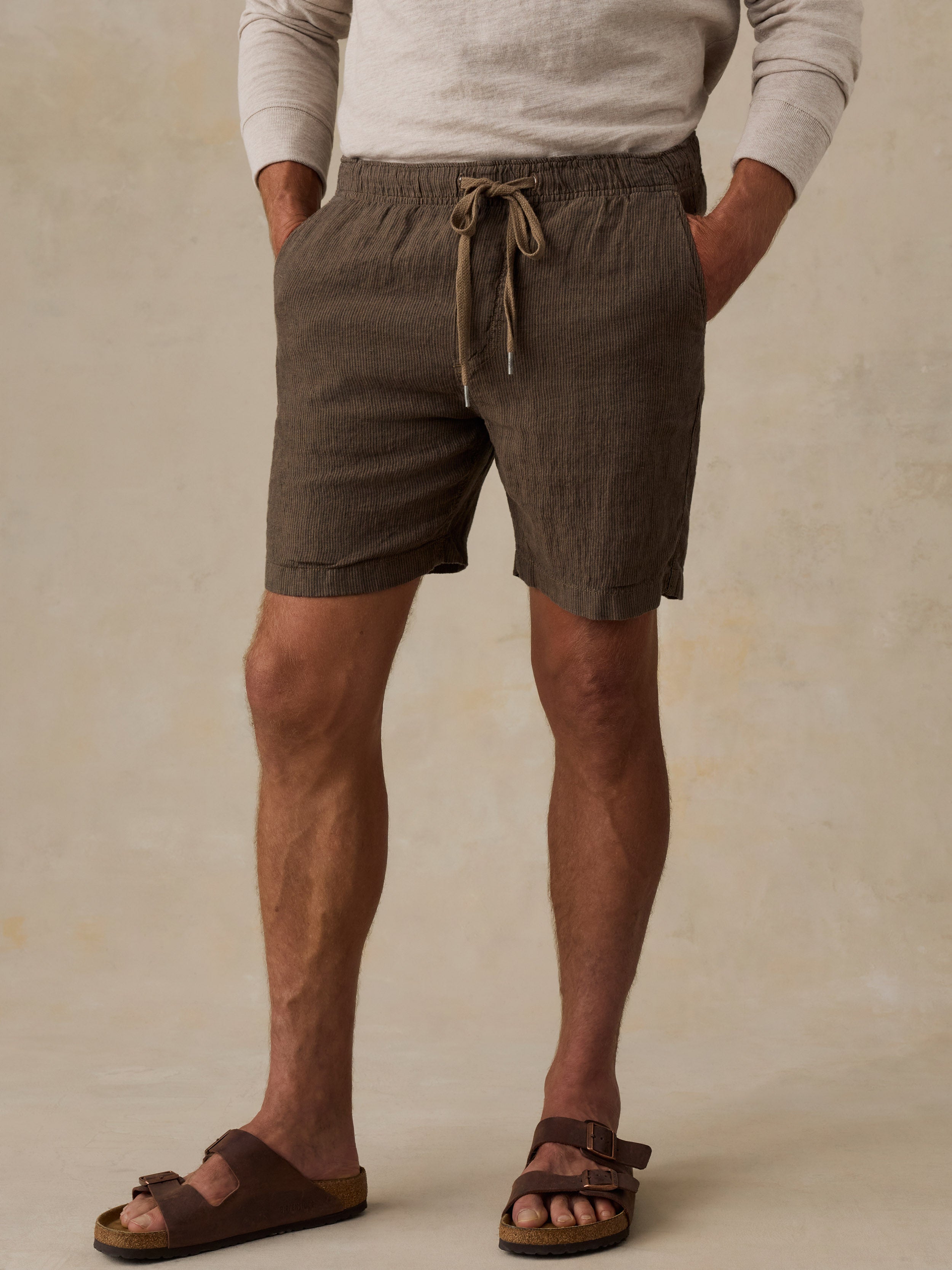 A man wearing dark brown linen drawstring shorts, a light beige long-sleeve shirt, and brown slide sandals, standing against a textured beige background.