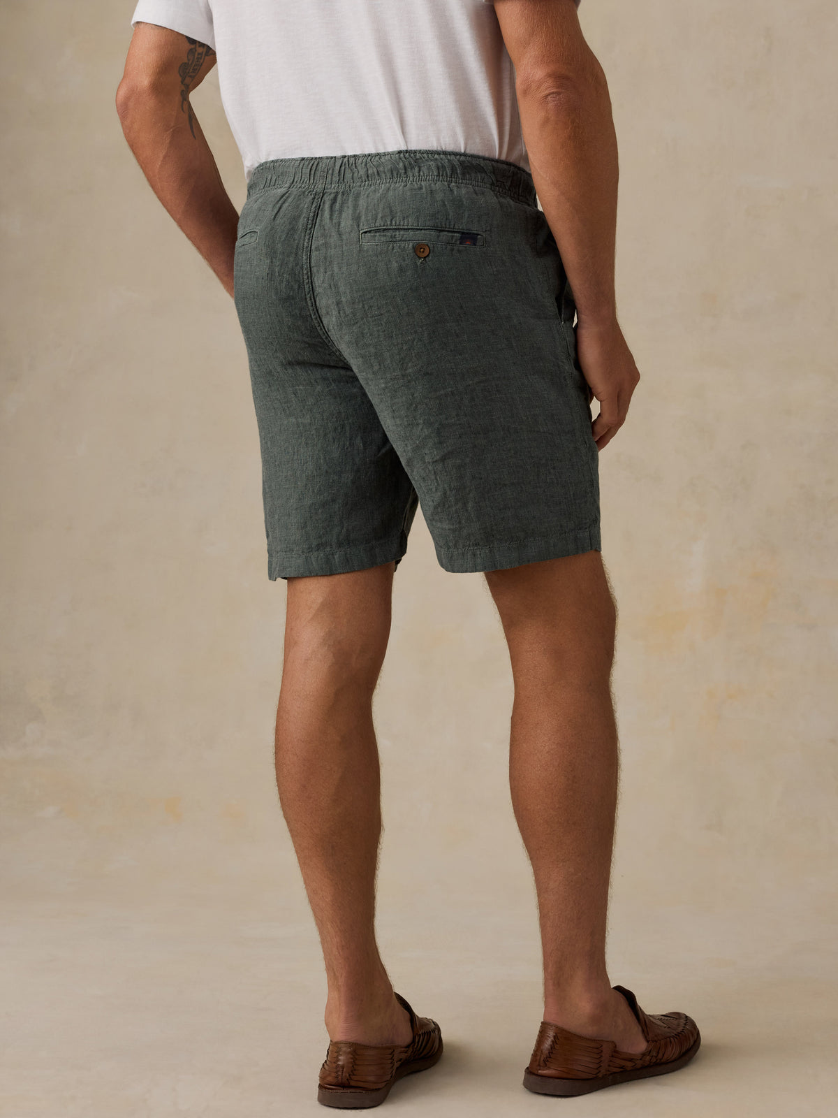 A man viewed from the back wearing dark green textured linen shorts, a white t-shirt, and brown leather loafers, set against a warm beige textured studio background., image 3 of 5