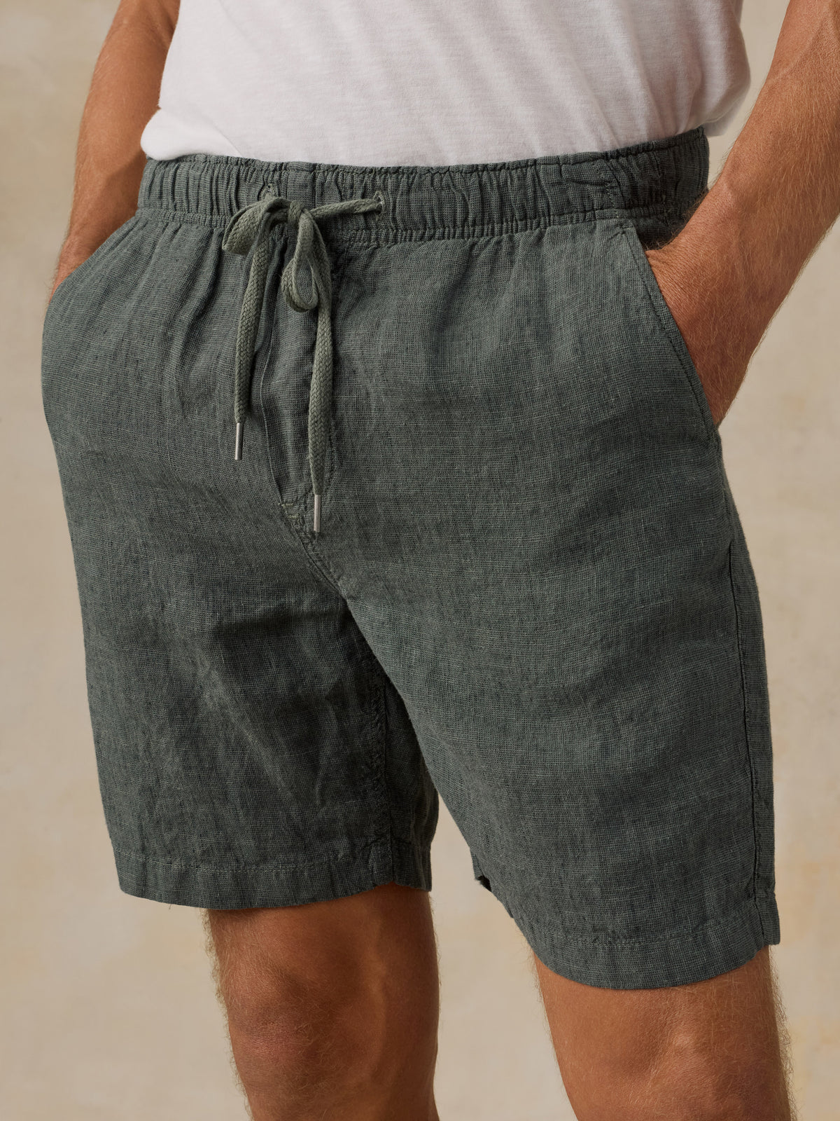 A close-up of a man wearing dark green drawstring shorts with a white t-shirt, standing against a neutral beige background., image 4 of 5